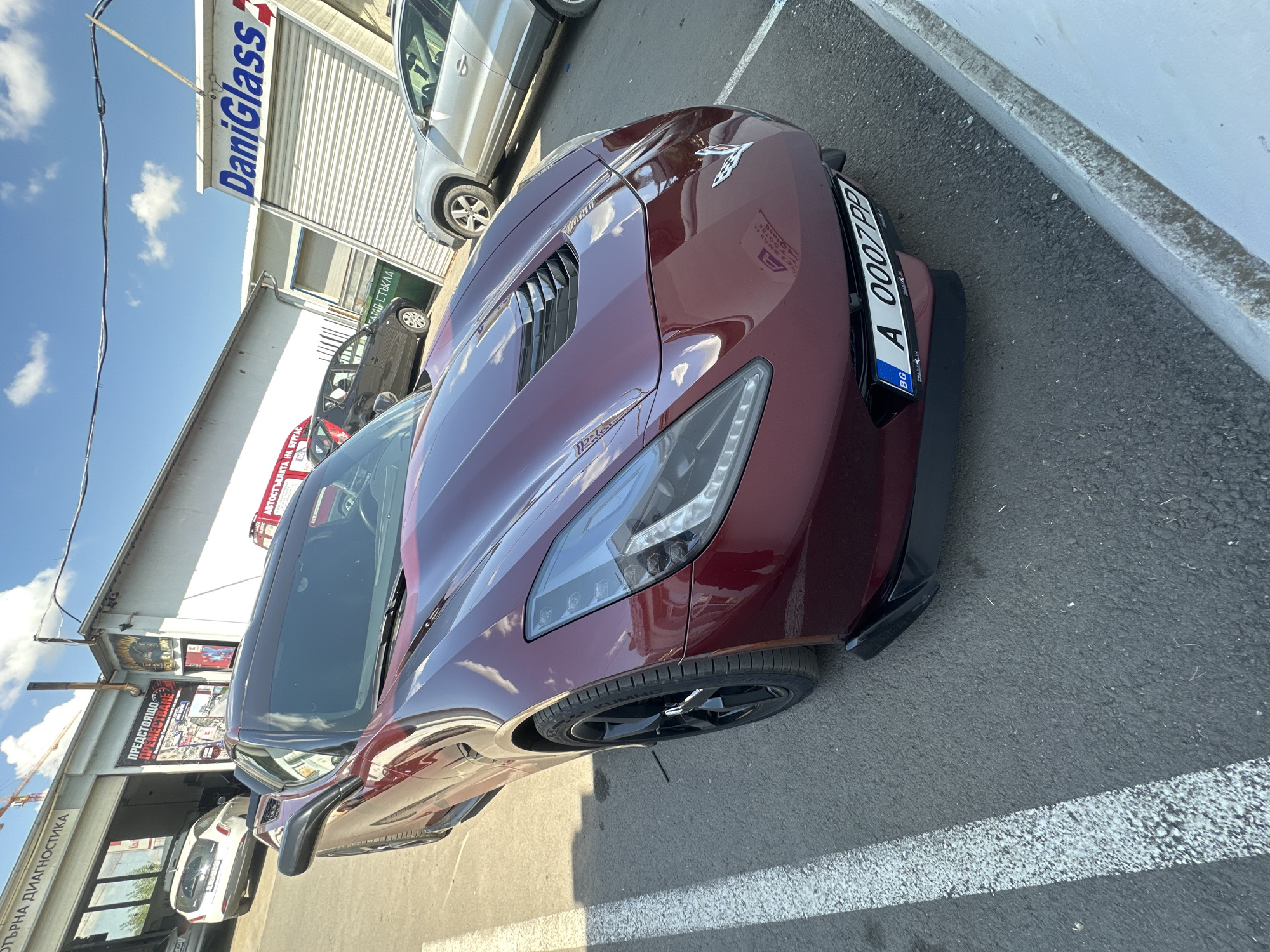 A red Chevrolet Corvette is parked in an outdoor lot. The car is facing the viewer at an angle. The car has a black spoiler on the front and black rims. The license plate reads "A 0007 PP". In the background, there are several buildings. One building has a sign that reads "DaniGlass". There are other cars parked in the lot, including a silver SUV and a white hatchback. The sky is blue with some clouds. The scene appears to be in an industrial or commercial area.