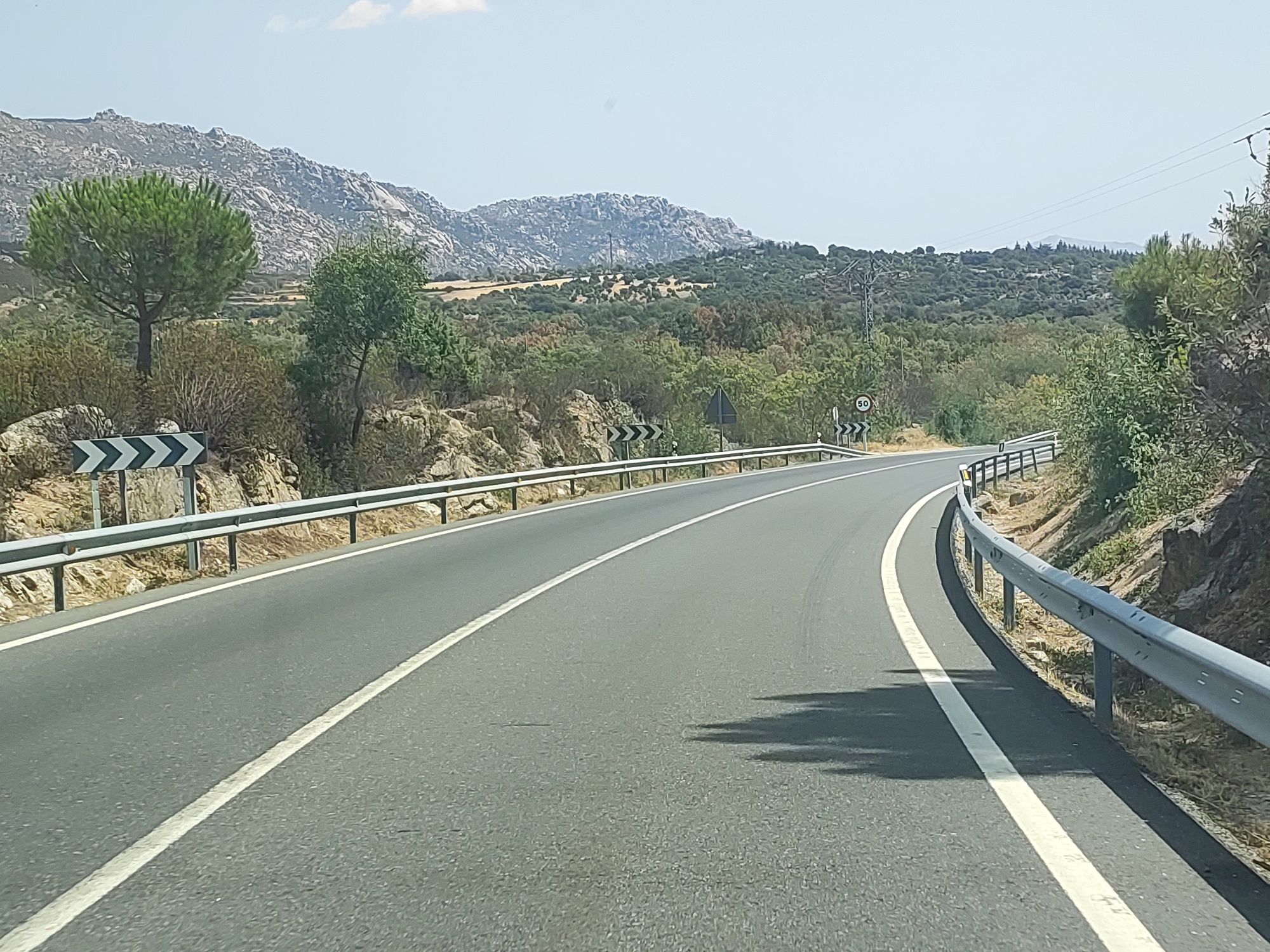 The image shows a two-lane asphalt road with white lane markings, curving to the right. A metal guardrail runs along the right side of the road, and another on the left side, separated by a shoulder and rocky terrain. The landscape features dry, rocky hills with sparse vegetation, including scattered trees and bushes. In the background, a mountain range with rugged peaks is visible under a clear, pale blue sky. A speed limit sign indicating "50" is mounted on a post near the guardrail on the right side of the road. The scene appears to be captured from a moving vehicle traveling along this road, likely in a rural or mountainous area. Given the context provided, this scene is located in Becerril de la Sierra, Spain.