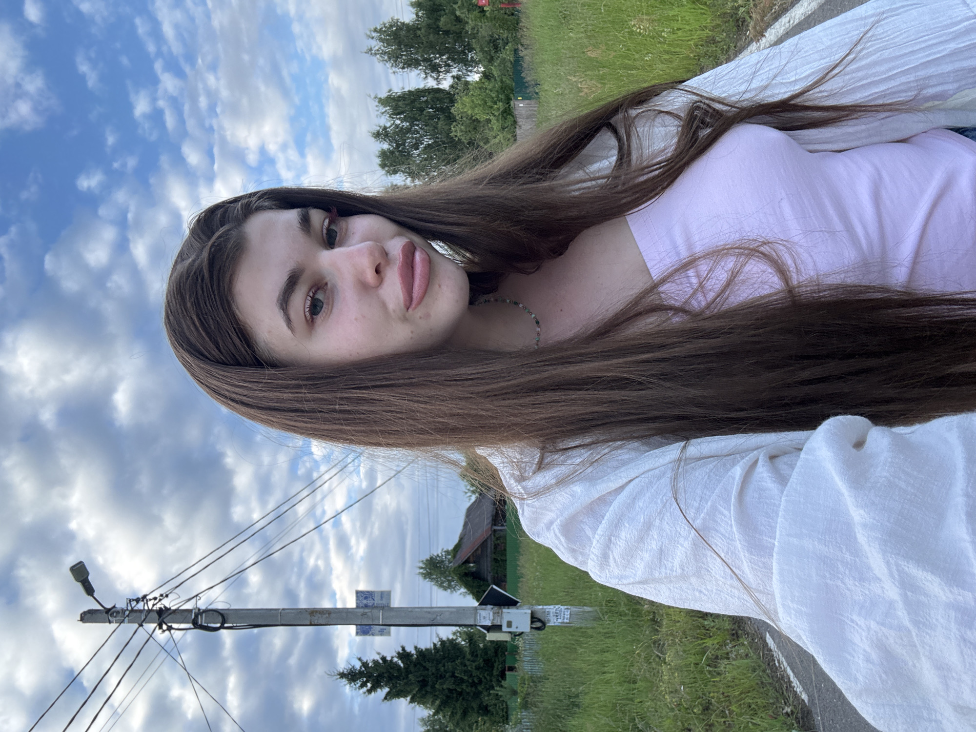 A young woman with long, dark brown hair is captured in a selfie, taken outdoors. She is wearing a light pink t-shirt and a white, textured garment draped over her shoulders. A beaded necklace is visible around her neck. Her expression is relaxed, with a slight smile and a gaze directed towards the camera.
The background suggests a rural or semi-rural setting. Tall green grass lines a paved road. In the distance, a rustic-looking wooden house with a dark roof is visible, nestled amongst trees. A utility pole with electrical wires and a streetlight stands prominently in the mid-ground, with a few small signs attached to it. The sky is filled with dramatic, fluffy white clouds against a bright blue backdrop, indicating a daytime scene with fair weather. The overall mood is casual and serene, possibly taken during a walk or a moment of relaxation outdoors.
The image appears to be taken in Мещёры, Russia, given the provided location context. There is no visible text within the frame.