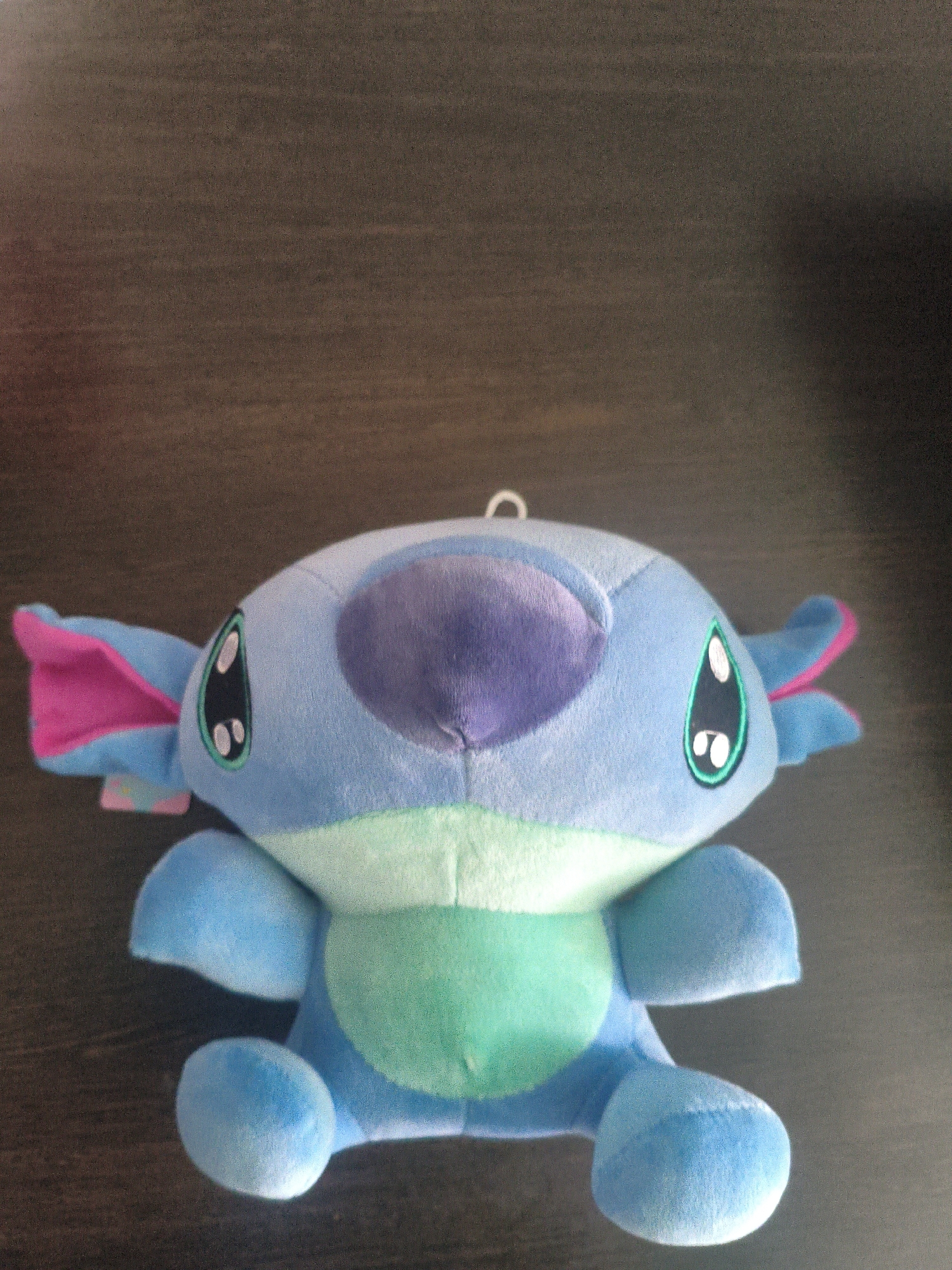 The image shows a blue plush toy resembling the character Stitch from Disney's Lilo & Stitch. The toy is positioned centrally on a dark brown wooden surface, likely a table. It has large, expressive eyes with black irises and white pupils, a purple nose, and pink and blue ears. The toy is seated with its arms and legs extended. The background is blurred and appears to be an indoor setting. The location of this scene in Lisakovsk, Kazakhstan, is not discernible from the provided image.