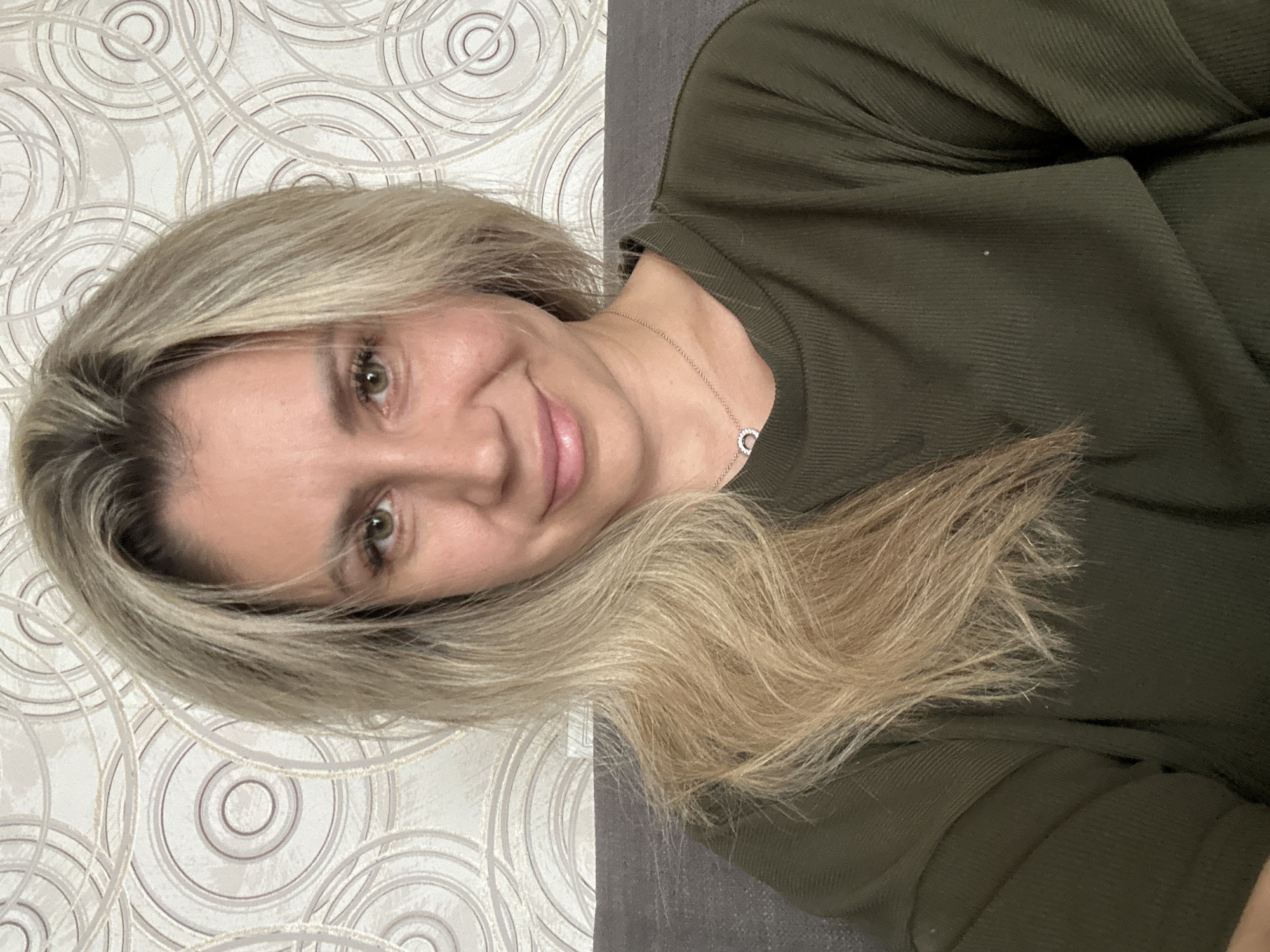 The image shows a close-up, head-and-shoulders selfie of a woman with blonde hair and green eyes. She is smiling and looking at the camera. She is wearing a dark green, long-sleeved shirt and a silver necklace with a circular pendant. The background is a textured wallpaper with a circular pattern. The image appears to have been taken indoors. There is no information in the image to indicate the location in the city of Novoaltaysk, Russia.