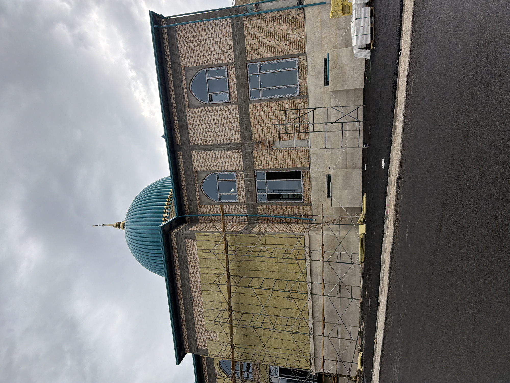 The image shows the exterior of a mosque under construction in Khumsan, Uzbekistan. The building features a large, teal-colored dome with a golden finial, and is constructed with light-colored bricks. Scaffolding surrounds a section of the building where yellow insulation is being installed. Various windows, some arched and some rectangular, are visible on the brick facade. The ground in front of the mosque is covered in fresh asphalt, and a pile of white tiles on pallets is present to the right. The sky is overcast with gray clouds.