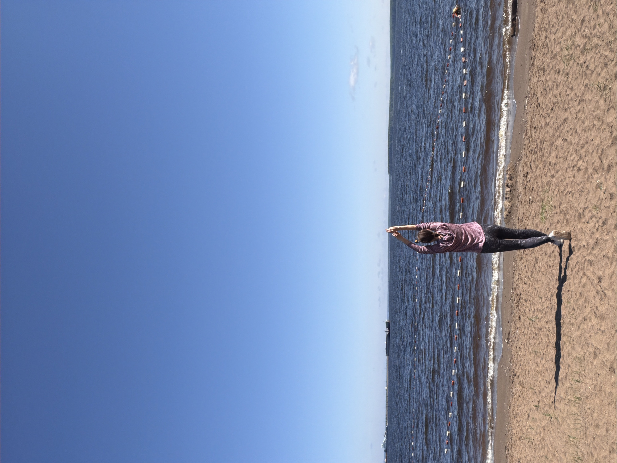 A person stands on a sandy beach with their arms raised above their head, facing the water. The person is wearing a pink hoodie and black leggings. The water is dark blue with small waves lapping at the shore. In the distance, there are several boats on the water, and a line of buoys separates the swimming area. The sky is clear and blue. The scene is the Kama River in the city of Chaikovsky, Russia.