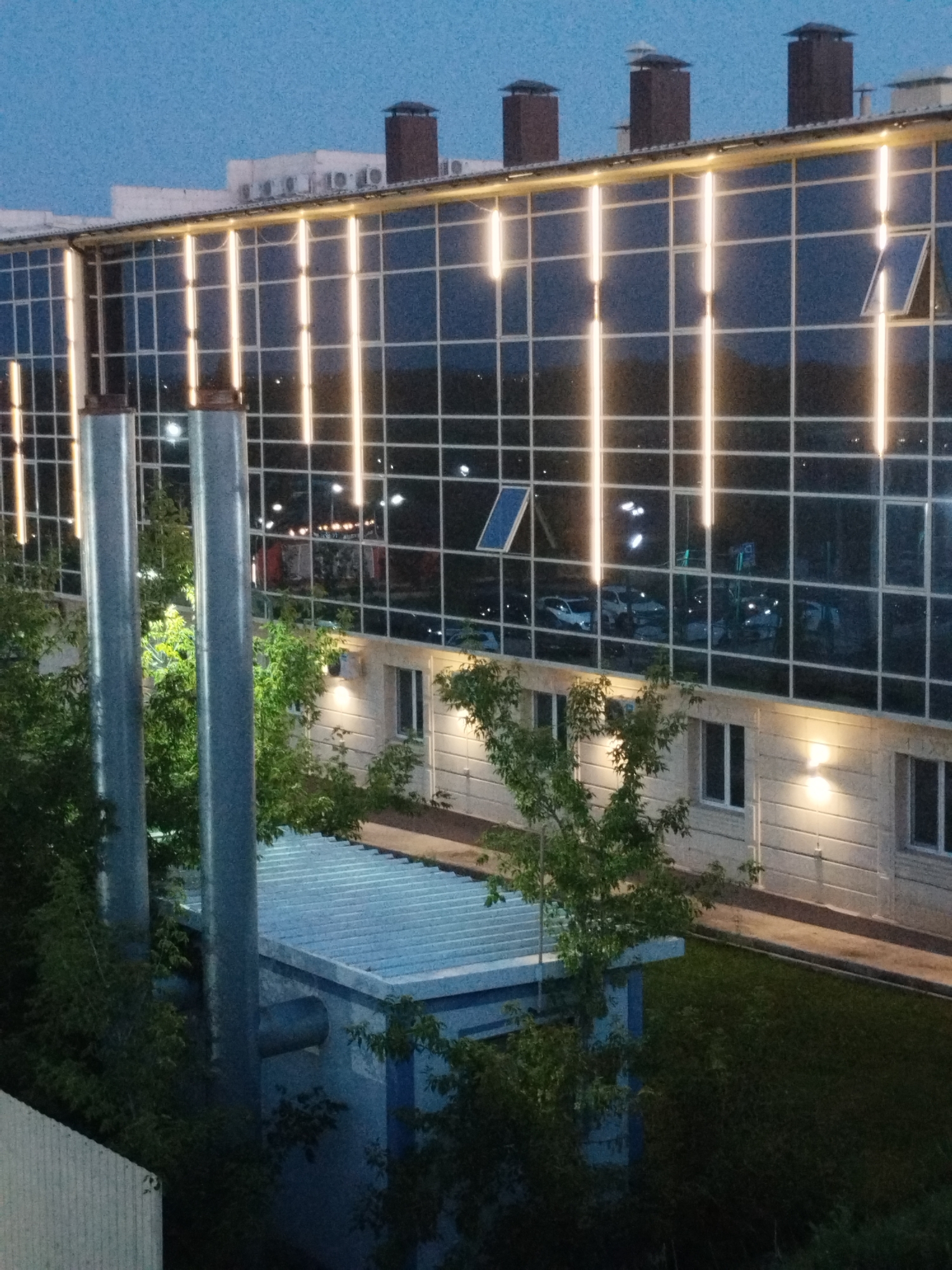The image captures an exterior view of a modern building at dusk in Kostanay, Kazakhstan. The building features a large, dark glass facade with vertical illuminated strips running across it, suggesting interior lighting or a decorative lighting system. Multiple windows are visible, some of which are open, revealing reflections of the exterior and possibly interior spaces. On the rooftop, several chimney-like structures are present. Below the glass facade is a section of the building with lighter colored siding and illuminated windows. In the foreground, several large, metallic cylindrical structures, possibly industrial pipes or vents, stand tall, partially obscured by lush green trees. A low-rise blue structure with a corrugated roof is visible to the right of the pipes, also surrounded by greenery. The overall environment appears to be an urban or semi-urban setting, given the presence of the building and the sky, which is a deep blue, indicating twilight. There are no people visible in the image.