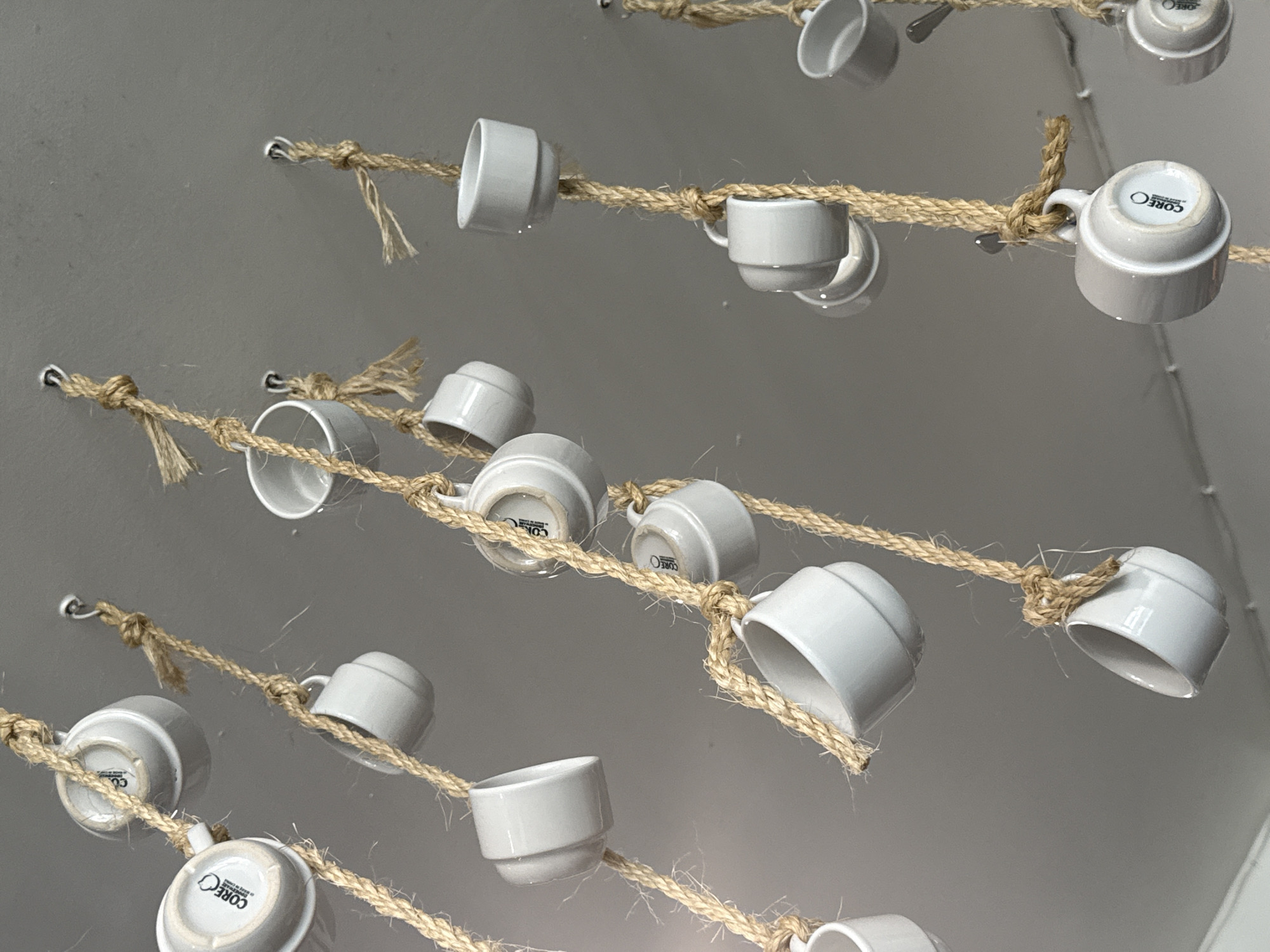 The image shows a ceiling-mounted art installation consisting of white ceramic cups strung on lengths of thick, natural-fiber rope. The ropes are secured to the ceiling with small metal hooks. The cups are spaced intermittently along the ropes, oriented in various directions, some with the "CORE" logo visible on their bases. The lighting is bright and even, suggesting an interior space. The background is a plain, light-colored wall or ceiling. The location of the photo is unknown.
