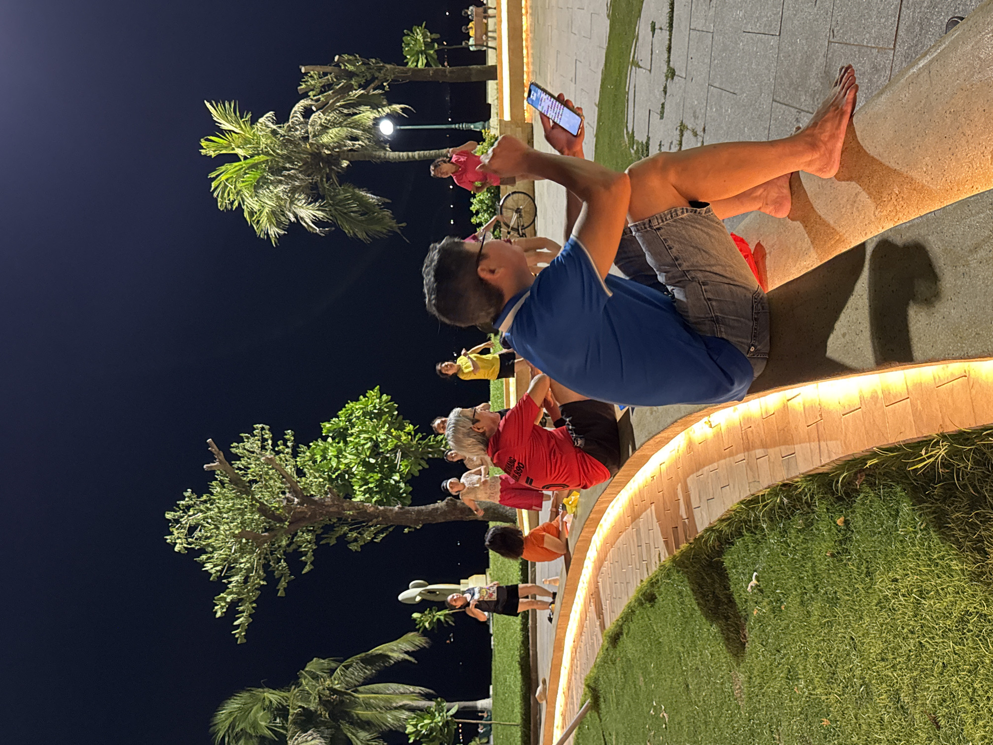 The image captures a nighttime scene in South Quy Nhon Ward, Vietnam, where a group of people are gathered outdoors. The setting appears to be a park or a public space with a paved walkway, lush green grass, and tropical palm trees. Several individuals are seated on a curved, illuminated concrete structure, suggesting a communal seating area.
One prominent subject is a young man in the foreground, wearing a blue shirt and denim shorts, holding a smartphone and looking at its screen. His bare feet are resting on the concrete. Behind him, several other people are visible, some sitting and others appearing to be engaged in an activity. One person in a red shirt is looking up at the sky, while another in a yellow shirt is also seated. Further back, a person in a pink top is standing, and a bicycle is parked nearby. The lighting suggests it's nighttime, with warm, artificial lights illuminating the pathway and seating area. The overall mood appears relaxed and casual, with people enjoying the evening.
Some text is visible on the red shirt of one of the individuals, but it is largely illegible due to the image quality and angle. The presence of palm trees and the general ambiance hint at a tropical coastal location.
