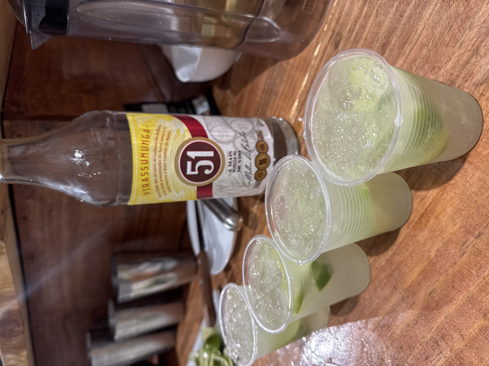 A bottle of "Cachaça 51" rests beside four plastic cups filled with a drink, likely caipirinhas, containing ice and lime wedges. The scene takes place on a wooden surface. The location is potentially Armação Dos Búzios, Brazil, given that cachaça and caipirinhas are common drinks in the region. The scene implies a social gathering or preparation for such, as multiple servings of the drink are ready.