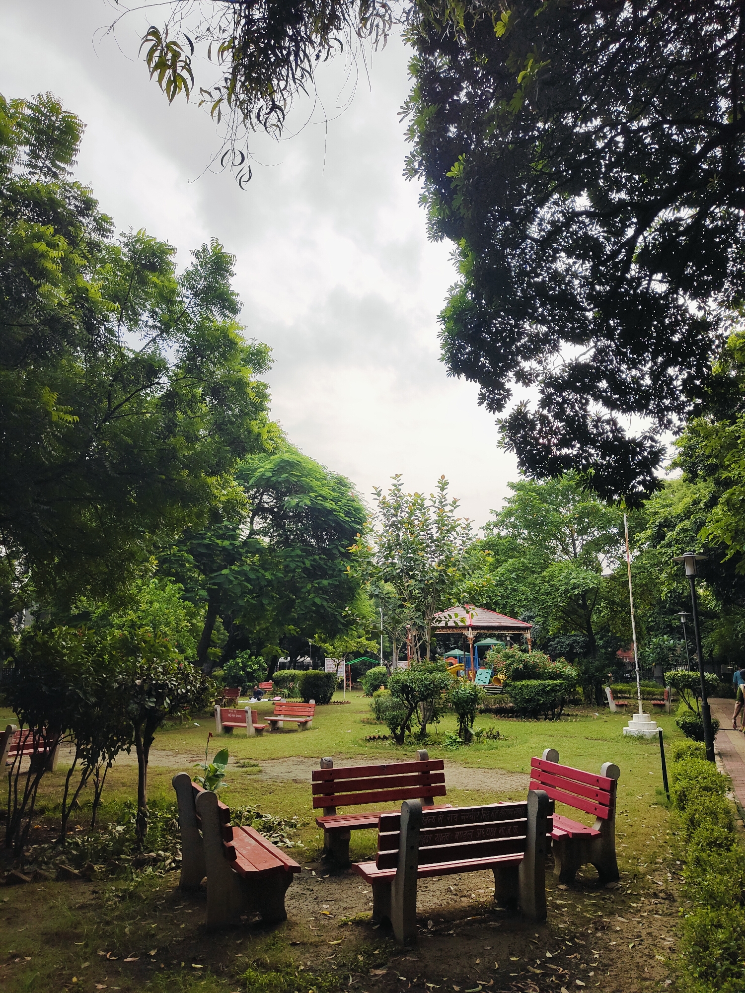 This is a photograph of a park in Gurgaon, India, on an overcast day. The park is filled with lush green trees and manicured bushes. Several red and concrete benches are scattered throughout the grassy area, with some positioned in the foreground and others further back. A playground with colorful structures is visible in the distance, under a pavilion. A paved walkway runs along the right side of the park, with a lamppost and a white pillar. A person can be seen walking on the walkway in the far right of the image. The sky is cloudy, contributing to a subdued lighting condition. Fallen leaves are scattered on the ground around the benches.