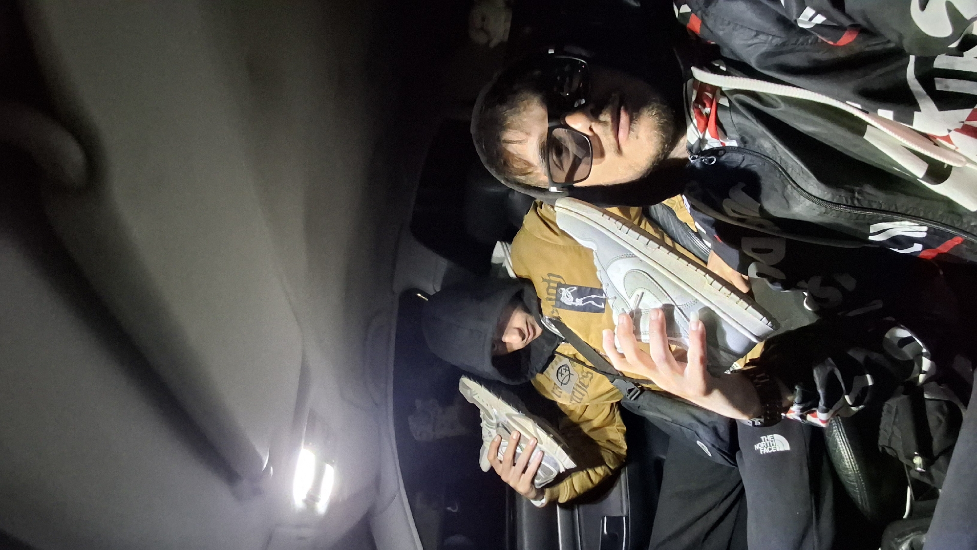 Two men are in the interior of a vehicle. One man is visible from the chest up and is wearing sunglasses and a black jacket with a white and red design. He is holding a gray sneaker in his left hand. Another man is hooded and wearing a yellow jacket with black detailing, and is holding a sneaker in his hands. The setting appears to be the inside of a car, possibly in Shumen, Bulgaria, though there is no specific evidence of this location within the image itself.