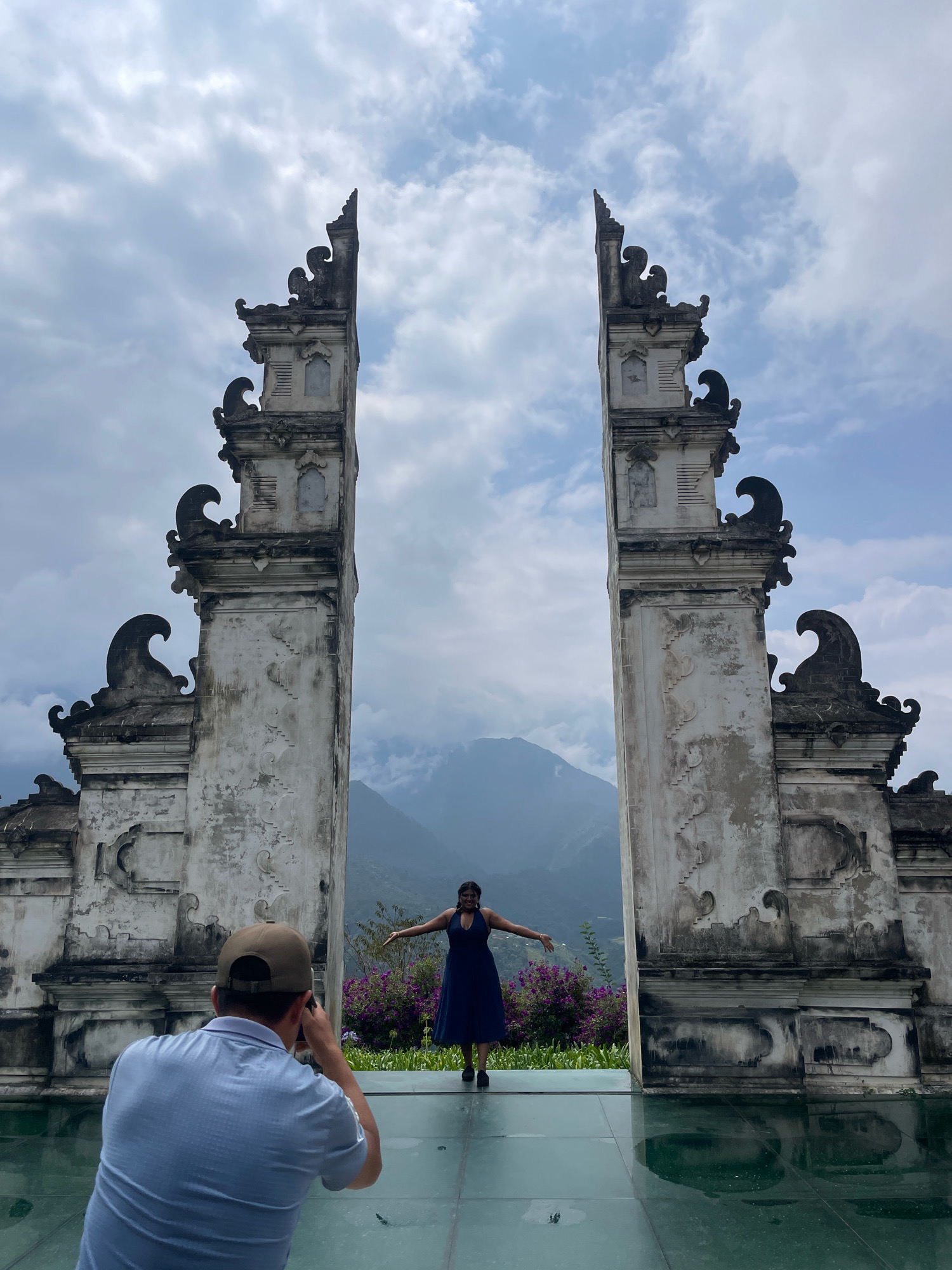 A medium shot captures a person being photographed in front of a monumental gate structure in Sa Pa Ward, Vietnam. The gate, constructed of weathered stone with ornate, carved details and a distinctive tiered roofline, stands symmetrically with two imposing pillars flanking a distant mountain range shrouded in clouds. In the foreground, a man wearing a light blue polo shirt and a tan baseball cap kneels, holding a camera and focusing on the person being photographed. The subject, a woman in a dark blue dress, stands with her arms outstretched, facing the camera. She is positioned on a transparent platform that reflects the sky and the gate structure. Behind her, a vibrant display of purple flowers adds a splash of color against the muted tones of the architecture and the misty mountain backdrop. The environment suggests an outdoor setting with a clear, albeit overcast, sky.