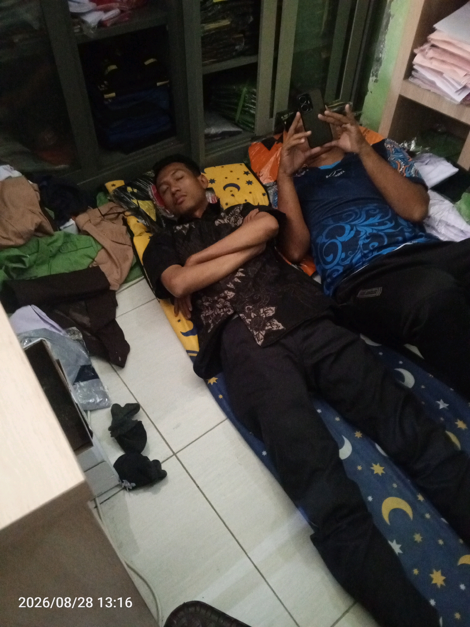 Two individuals are lying on the floor of what appears to be a room with tiled flooring. The room contains shelves with stacked items and clothing. In the foreground, one person is lying on their back with their arms crossed, wearing a dark patterned shirt and dark pants. Next to them, another person is lying on their side, holding a black smartphone up to their face. They are wearing a blue t-shirt with a white pattern and black pants. There are various pieces of clothing and bedding scattered around the individuals. The timestamp "2026/08/28 13:16" is visible in the bottom left corner of the image. The location is identified as Randegansari, Indonesia.