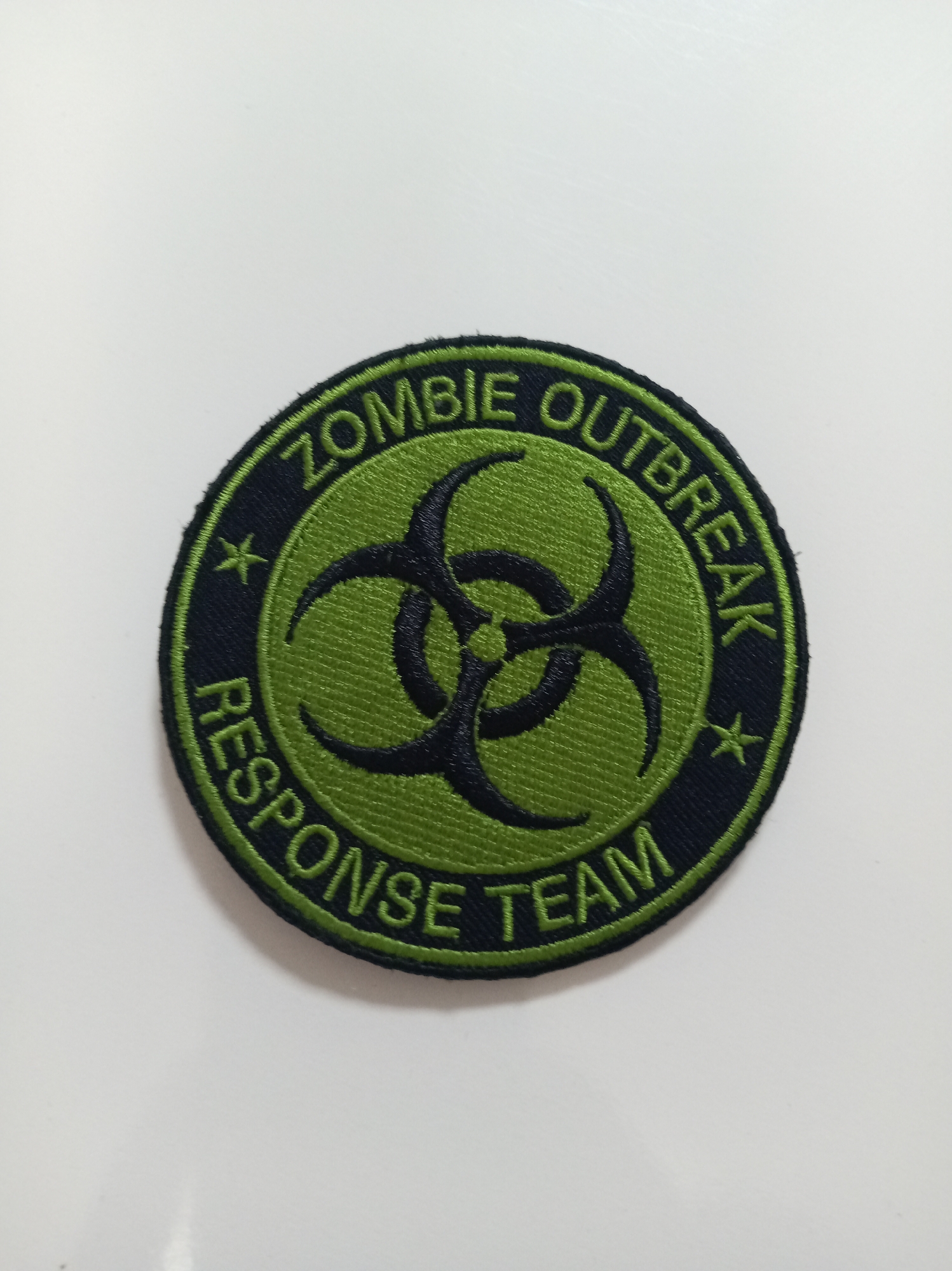 A circular embroidered patch with a biohazard symbol in black on a green background is centered against a white surface. The patch features text in green embroidered letters that reads "ZOMBIE OUTBREAK RESPONSE TEAM" around the biohazard symbol. Small black stars are also present on the patch.