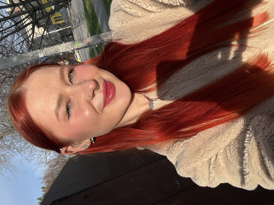 The image displays an outdoor, close-up portrait of a female individual located in Rostov-on-Don, Russia. The subject has long, bright red hair and fair skin with visible freckles across her nose and cheeks. Her eyes are green, and she presents a subtle smile with pink-glossed lips, looking directly into the camera. She wears a silver horseshoe septum piercing, a small silver stud in her right nostril, and multiple silver piercings in her visible left ear, including hoops and studs. Her attire consists of a light beige, zippered fleece jacket worn over a darker, possibly grey, undergarment. The scene is illuminated by strong sunlight, casting distinct shadows on her hair and jacket. The background on the left features a dark, corrugated metal fence or structure. To the right, a city street is visible with a utility pole adorned with stickers, bare trees, a strip of green grass, a multi-story building with a light-colored facade, and a white vehicle with a yellow stripe, likely a taxi, parked further down the street.