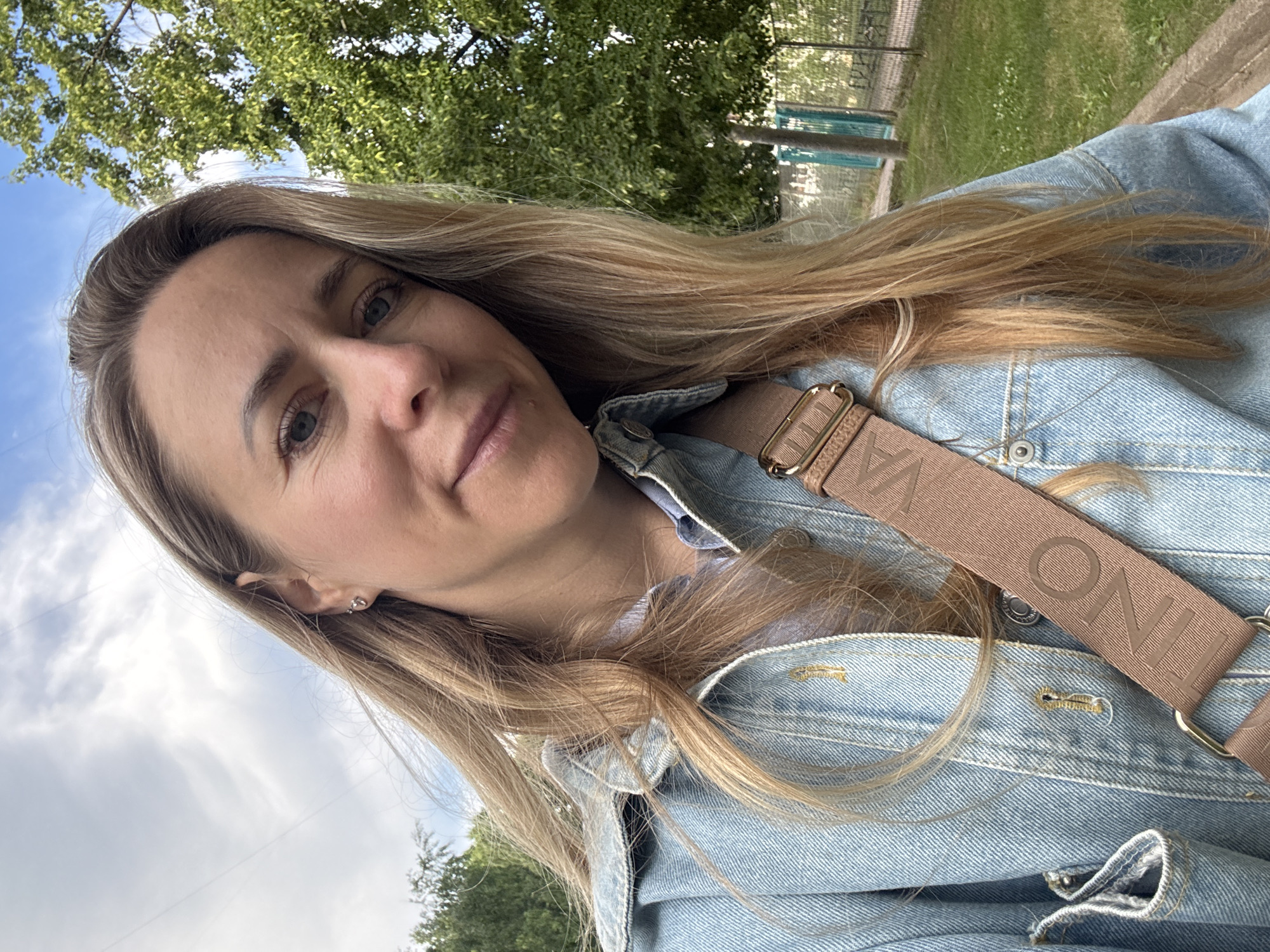 A woman with blonde hair, wearing a light blue denim jacket over a blue collared shirt and a tan strap with the word "VALENTINO" printed on it, takes a selfie outdoors. She has blue eyes and is looking at the camera with a slight smile. She is wearing a small stud earring in her left ear. The background shows a cloudy sky, green trees, and a fence. The scene appears to be in a park or outdoor recreational area.