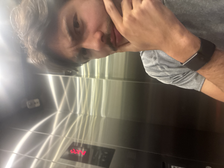The image displays a male individual, appearing to be in his 20s or 30s, inside a polished metal elevator. The subject occupies the right half of the frame, facing the camera directly, suggesting a selfie. He has dark, wavy hair, a mustache, and a short beard. His expression is one of contemplation or slight puzzlement, with a furrowed brow and his right hand (from the viewer's perspective) resting with fingers on his chin. He is wearing a grey short-sleeved t-shirt and a dark fitness tracker or smart band on his left wrist. The elevator interior features reflective stainless steel walls, showcasing multiple bright, linear reflections of overhead lighting. On the left wall, a digital display indicates the number "2" in red, signifying the current floor, with control buttons visible beneath it. A small, light-colored security camera is mounted in the upper-left corner of the elevator cab. The specific location within the city of Mexico City, Mexico, cannot be determined from the visual information provided within the image.