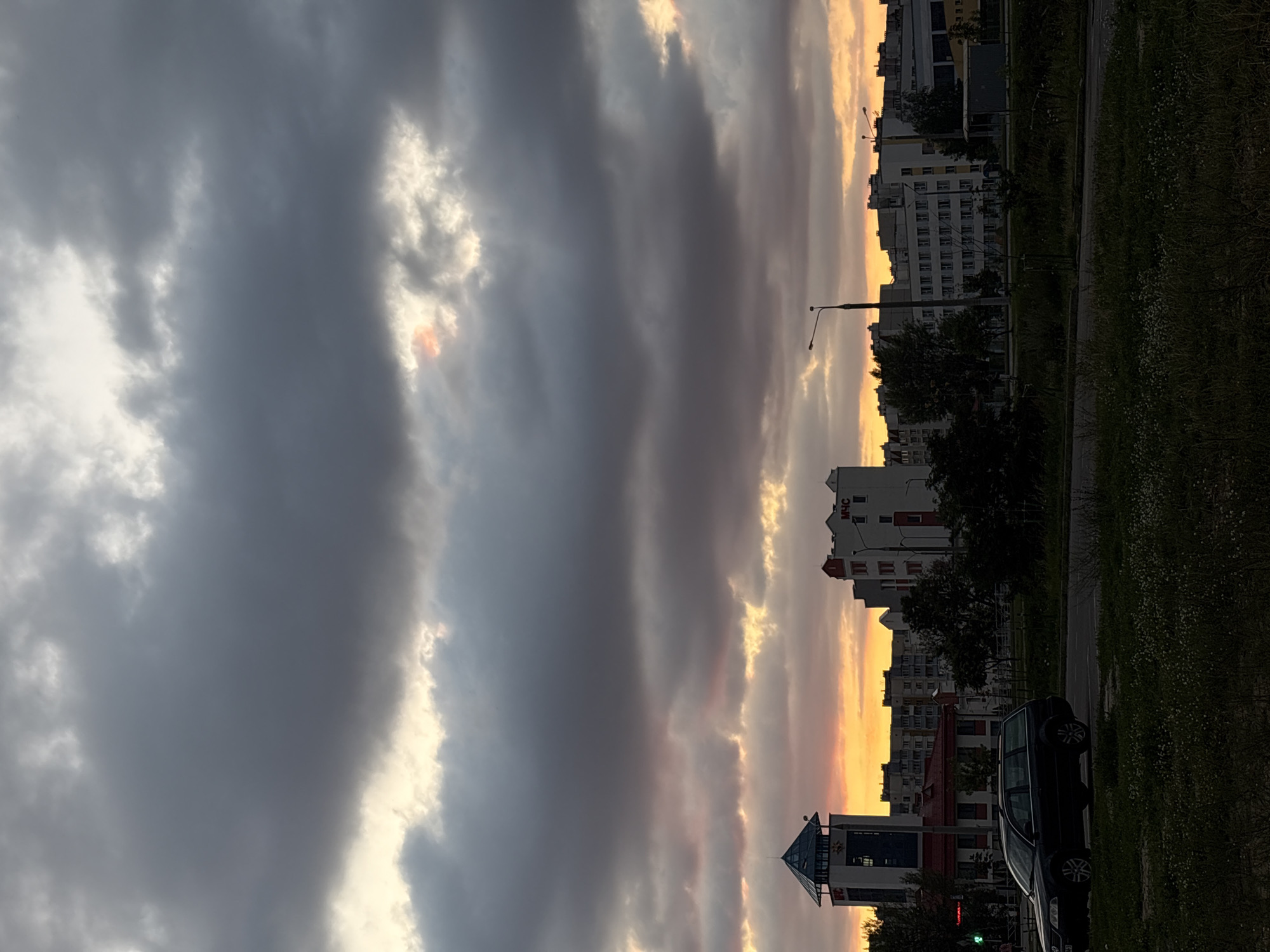 The image shows a street scene in the city of Unknown during sunset. The sky is filled with dark clouds, with patches of orange and yellow light peeking through, indicating the setting sun. In the foreground, a dark-colored station wagon is parked on the right side of a road. The road appears to be a multi-lane highway, with some grass and vegetation along the median and sides. In the background, a series of apartment buildings and other structures are visible. One prominent building on the left has a blue, pyramid-shaped roof and a sign with "MFC" written on it. Another building on the right is white with red accents and has the Cyrillic letters "МЧС" on it. There are streetlights visible along the road. The overall atmosphere of the image is one of a city winding down as the day comes to a close.