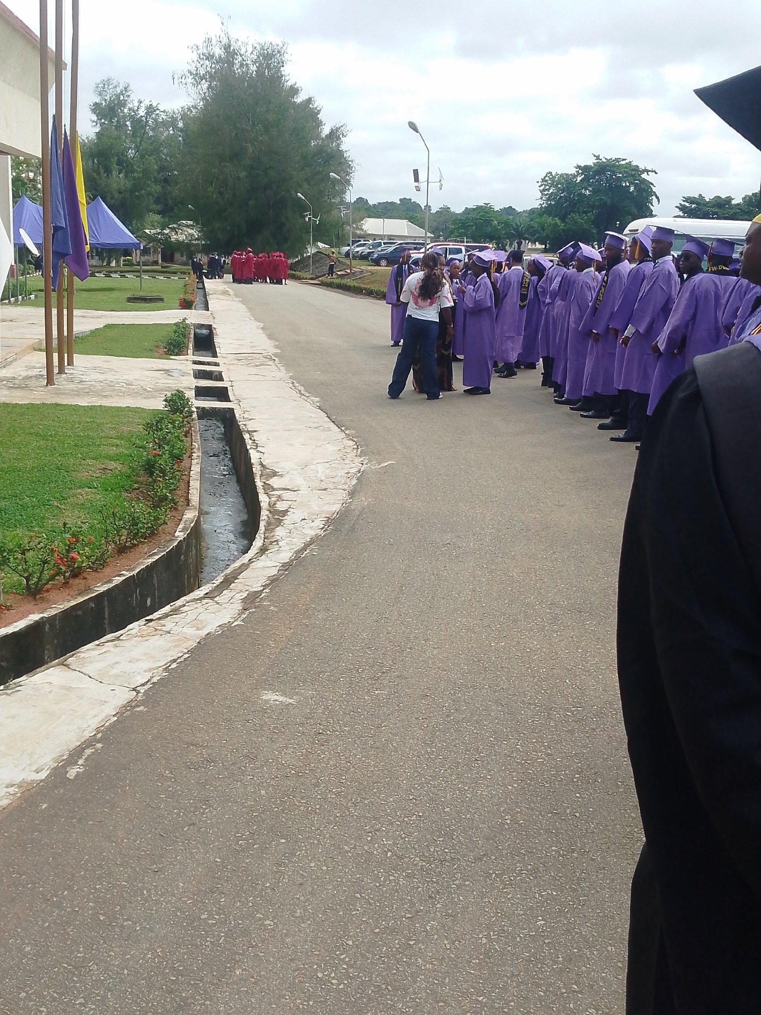 A group of graduates, all wearing purple academic gowns and caps, are lined up on a paved walkway next to a grassy area and a drainage ditch. Some are looking towards the camera, while others are looking forward. One graduate in the front row is looking at a person who appears to be addressing them. In the background, there are trees, buildings, and cars, suggesting an outdoor campus setting. The sky is overcast, indicating a cloudy day. The scene appears to be a graduation ceremony. There is no visible text.