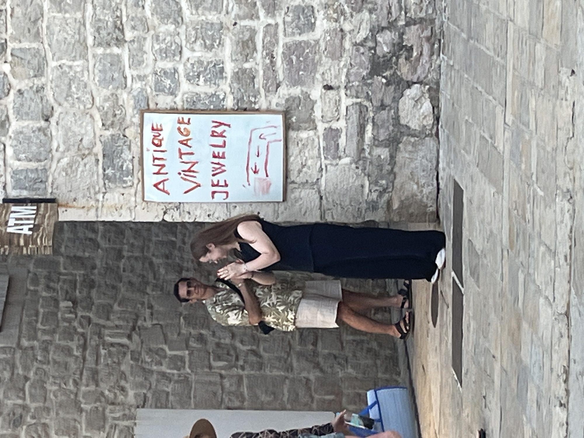 The image depicts a street scene in Budva, Montenegro, with a focus on a stone wall and a sign. A man wearing sunglasses, a patterned short-sleeved shirt, and light-colored shorts stands on the left, facing right. He has a dark bag slung across his chest. To his right, a woman in a black sleeveless top and wide-leg pants faces the man, her hands clasped together. A white sign with red lettering, reading "ANTIQUE VINTAGE JEWELRY" and a simple arrow diagram, is displayed on the stone wall. The wall is constructed of rough, grey stones. A portion of an ATM sign is visible at the top left of the frame. The ground is paved with stone tiles. The overall lighting suggests it is daytime, possibly late afternoon given the shadows.