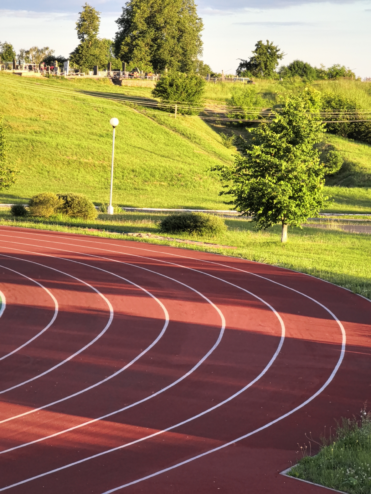 A low-angle outdoor photograph captures a portion of a red running track with white lane lines. The track curves towards the right of the frame. Beyond the track, a grassy incline rises, dotted with several small bushes. In the middle ground, a single street lamp stands on a metal pole. Further up the hill, a cemetery is visible with numerous gravestones and monuments. Several trees, including some with dense green foliage, are scattered across the hillside and in the distance. Power lines are strung across the grassy slope. The sky above is a pale blue with some scattered clouds. The scene is illuminated by the warm light of what appears to be late afternoon or early morning sun, casting long shadows across the track. There are no people or animals present in the image.