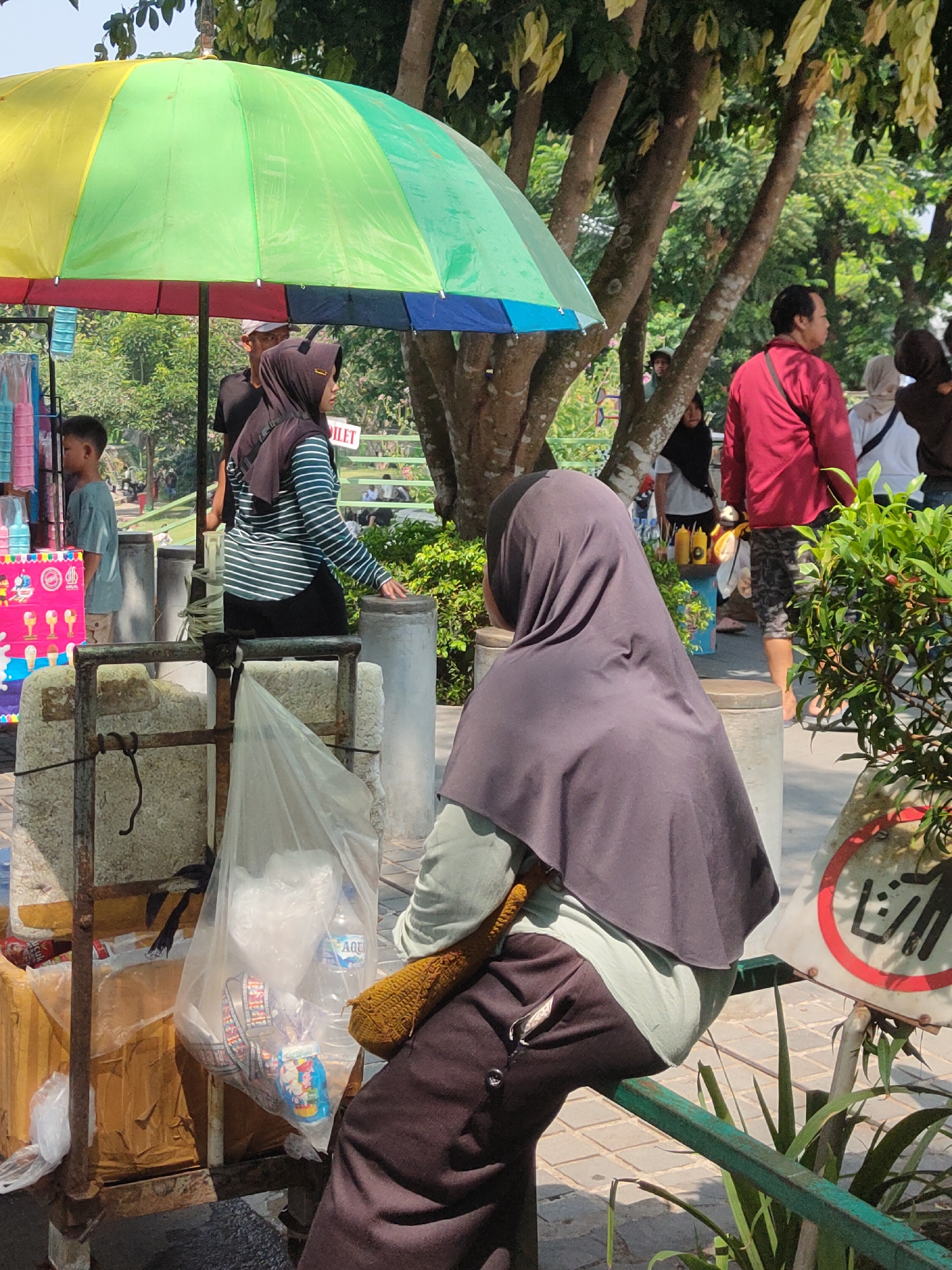 In the city of Ngasem, Indonesia, under a vibrant yellow, green, and red umbrella, a street vendor displays colorful candies and drinks on a cart. A young boy in a light blue t-shirt stands nearby. Two women wearing hijabs are present; one in a striped shirt is by the cart, and another in a dark hijab is seated. Behind them, a man in a red shirt walks away, and other individuals are visible in the background. The scene is set outdoors with trees providing shade. A sign depicting a "no littering" symbol with Japanese characters is visible on the right.