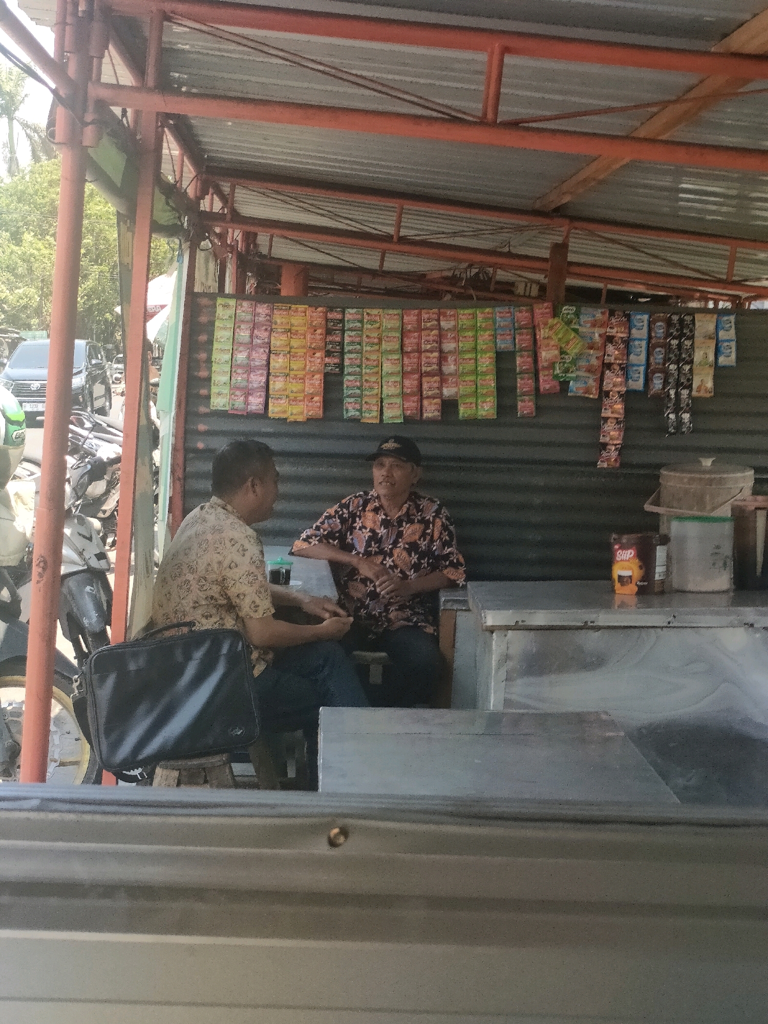 Two men are sitting at a stall in Waru, Indonesia. The stall is open to the street and has a metal roof supported by orange metal beams. The back wall of the stall is corrugated metal, and a variety of packaged goods are displayed on racks above the counter. The man on the left is wearing a patterned shirt and dark pants and is looking away from the camera. The man on the right is wearing a dark patterned shirt and a black baseball cap and is facing the man on the left. They appear to be engaged in conversation. In front of the men is a counter with various items, including a cup of coffee, a container, and a product labeled "GULAI". To the left of the stall, motorcycles are parked, and a car is visible in the background, suggesting a busy street. The overall lighting suggests it is daytime.