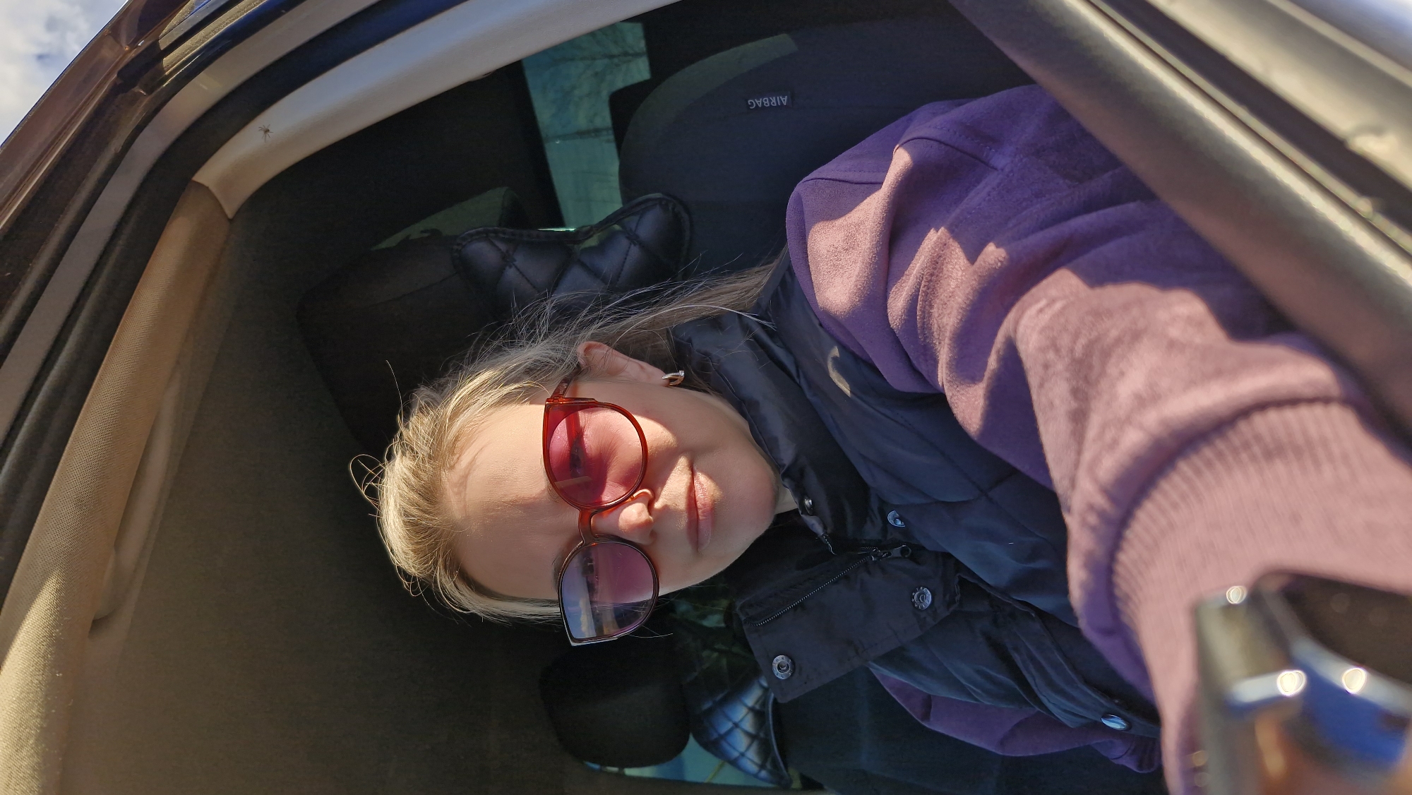 The image displays a young woman inside a car, positioned diagonally across the frame. She has blonde hair and is wearing red-framed sunglasses, a purple hooded sweatshirt, and a black quilted vest. Her expression is neutral, and her eyes are obscured by the sunglasses, which reflect bright light. Part of a shiny metallic object, likely a mobile phone being used to take the selfie, is visible in the bottom right corner.
The setting is the interior of a car, with the dark headrest of a car seat visible behind the woman's head. The word "AIRBAG" is printed upside down on this headrest. Above and behind her, the car's ceiling, a side pillar, and part of the window frame are visible, with bright sunlight streaming in. A small, light-colored spider can be seen clinging to the car's interior trim near the top-left corner of the image.
The strong sunlight indicates it is daytime, casting prominent shadows across her clothing and face. The scene suggests the woman is either relaxing or traveling in the car in Otchor, Russia. The image itself appears to be a rotated selfie.