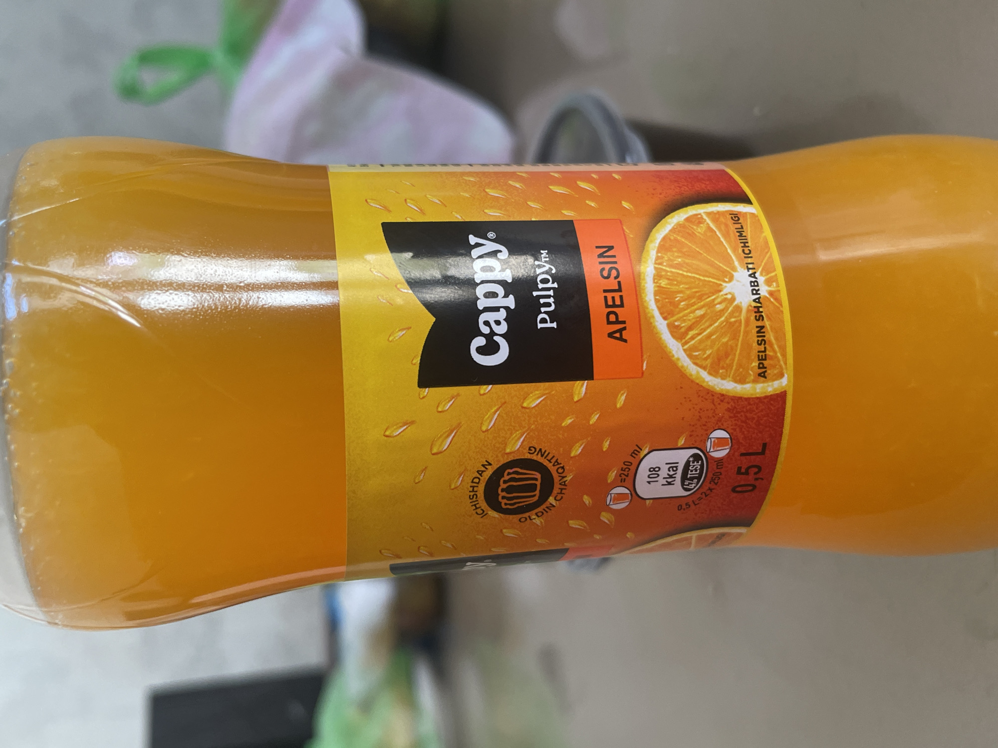 A close-up, eye-level shot focuses on the label of a plastic bottle filled with orange juice. The bottle is positioned centrally, with the label taking up most of the frame. The liquid inside is a vibrant orange, with small bubbles visible near the top.
The label is predominantly yellow and orange, featuring water droplets scattered across its surface. Prominently displayed is the brand name "Cappy" in white, outlined in black, followed by "Pulpy™" in smaller white lettering. Below this, a red rectangular band contains the word "APELSIN" in white, indicating the flavor. An image of a sliced orange is depicted on the lower right portion of the label, with text in a smaller font curving around it, reading "APELSIN SHARBATI ICHIMLIGI".
To the left of the "Cappy" logo, there's a circular emblem with text in an unfamiliar script, possibly featuring a stylized illustration of crowns or cups. Below this, nutritional information is presented, showing "≈250 ml", "108 kkal", and "4% TESE+". Further down, the volume "0,5 L" is clearly visible. A smaller text underneath reads "0,5 L = 2 x 250 ml".
The background is blurred, suggesting a shallow depth of field. On the left, a dark rectangular object, possibly a laptop or monitor, is partially visible. To the right, a light-colored surface with some pink and white fabric, and a hint of a green plastic bag, are discernible. In the lower right background, a dark grey, opaque container or lid is also visible.
The lighting is soft, with some highlights reflecting off the plastic bottle and the glossy label. The overall impression is that of a product shot, likely taken indoors, perhaps in a kitchen or a commercial setting. The image does not provide any specific visual cues to pinpoint its location within the city of Tashkent, Uzbekistan.