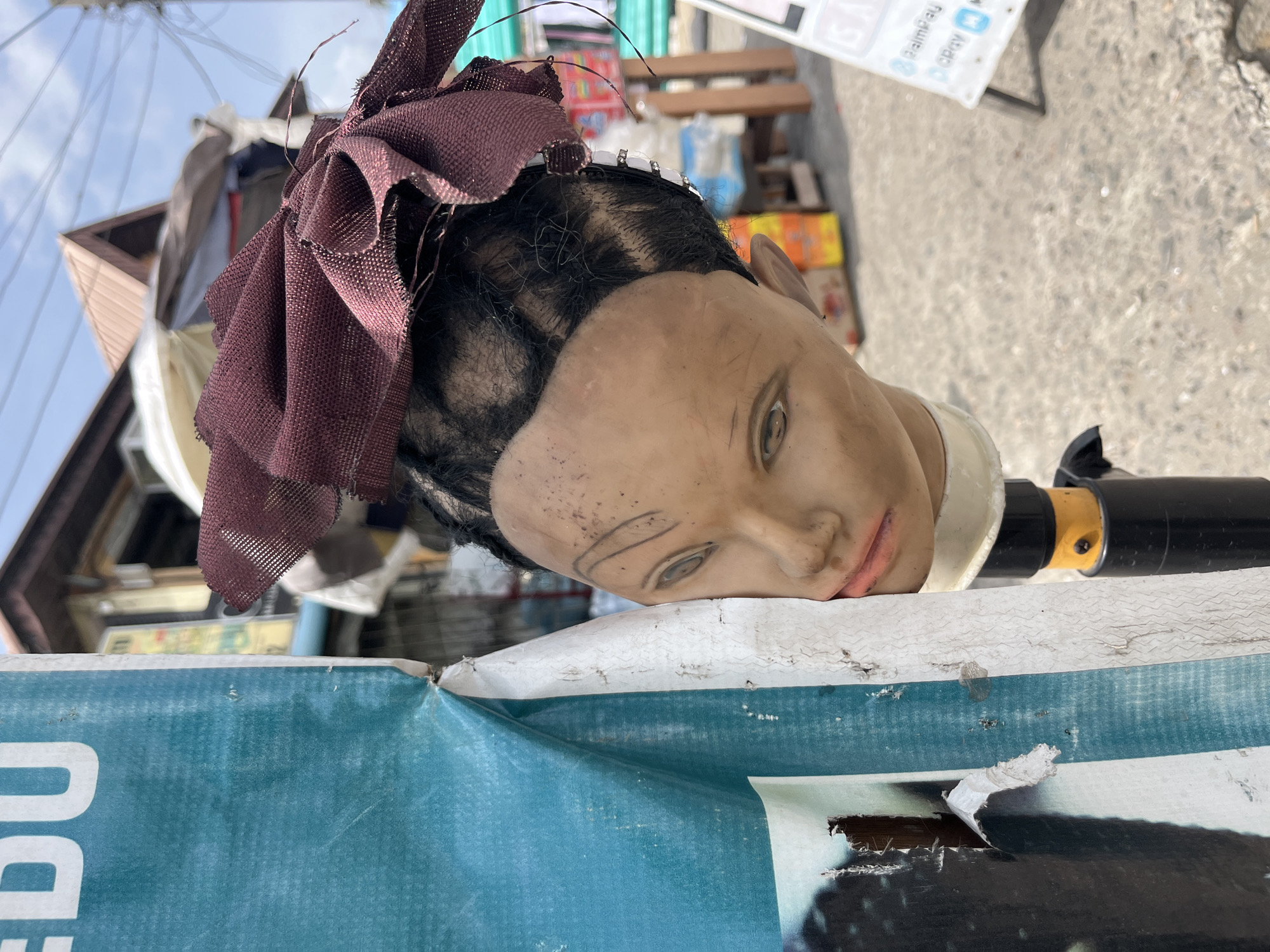 The image displays a mannequin head resting on its right side atop a folded, worn sign outdoors. The mannequin head, appearing to be made of plastic or a similar material, has a light skin tone with dark, short hair. A dark maroon or brown fabric, tied in a bow, is positioned on top of the head, acting as a headscarf or hair accessory. A thin white headband with small, clear embellishments is visible near the forehead. The face has drawn-on eyebrows and eyes that gaze slightly downward and to the right, with a hint of a pinkish tint on the lips. Specks of dirt or dust are noticeable on the mannequin's forehead.
The mannequin head rests on a large, heavily worn banner or sign, predominantly teal/turquoise in color with a white band. The sign shows significant damage, with rips and peeling revealing underlying layers. Partially visible white text on the sign includes "DU" and "edu". To the right of the mannequin's neck, a black object with a yellow ring is partially visible, possibly part of a stand.
The background, under a bright sky, suggests a daytime outdoor setting. In the upper left, parts of buildings or structures with brown roofing and light-colored siding are visible, crisscrossed by multiple utility wires. The upper right background shows a blurry, grey, textured ground surface. Another blurry white sign in the distance displays the text "PalmPay" and "opay". The scene is static, depicting an unattended mannequin head in an urban or semi-urban environment, consistent with Ado, Nigeria.