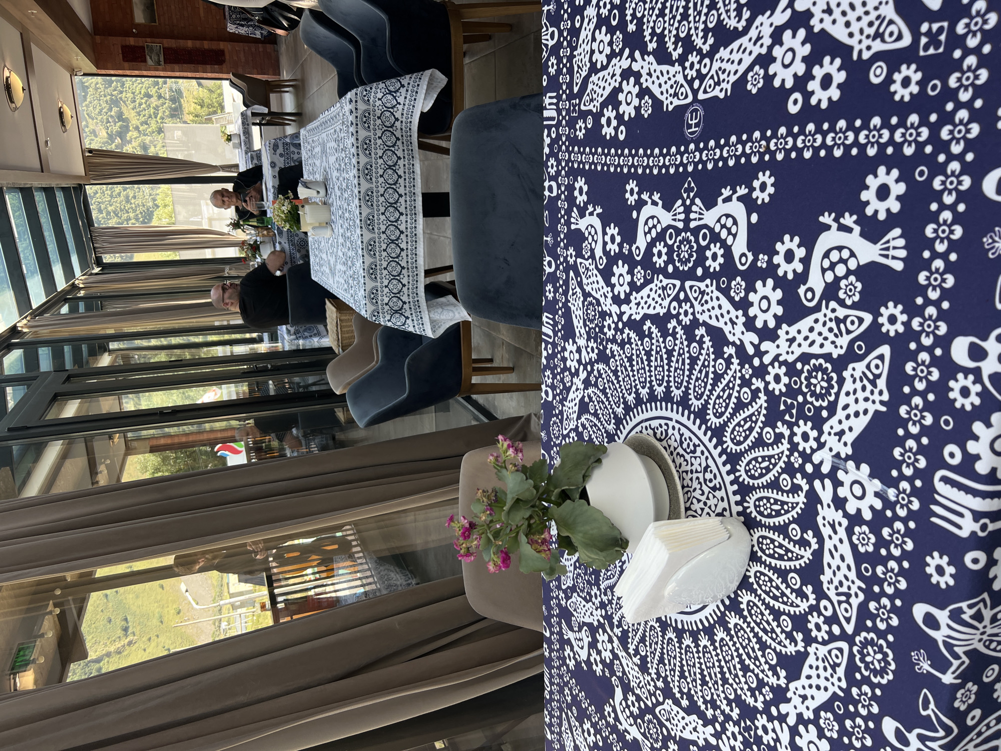 The image depicts the interior of a restaurant in Stepantsminda, Georgia, with a focus on a table in the foreground covered with a dark blue tablecloth featuring a white paisley and folk-art pattern of birds and flowers. On this table, a white ceramic pot contains a small flowering plant with pink blossoms. A white napkin holder filled with white napkins is positioned to the left of the plant. In the background, through a large window, a mountainous landscape with green slopes and trees is visible under a bright sky. Two men are seated at a table in the middle ground, which is draped with a white tablecloth patterned with blue fish and geometric designs. They are facing away from the camera, engaged in conversation. The restaurant appears to have a modern design with a glass roof and large windows, offering a view of the natural surroundings.