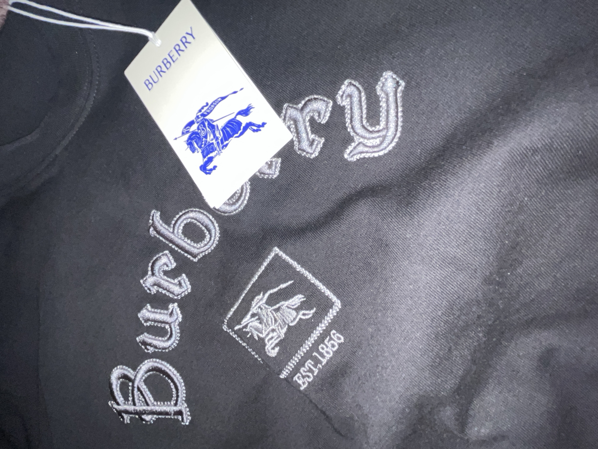 The image presents a close-up view of a black garment, likely a sweatshirt or hoodie, adorned with distinctive embroidery and an attached brand tag. The fabric of the garment exhibits a subtle diagonal texture.
Prominently featured on the garment is silver-grey embroidery. The main text, "Burberry," is stitched in a decorative, gothic-style font. Below this, a square-shaped embroidery contains the iconic Burberry Equestrian Knight logo, with the text "EST.1856" positioned to its right. The embroidery is detailed and appears to have a raised, textured quality.
A white brand tag, connected by a thin white string, is attached to the garment. The tag clearly displays "BURBERRY" in bold blue capital letters. Below the brand name, the blue Burberry Equestrian Knight logo is printed, accompanied by the word "PRORSUM" near the horse and rider figure.
The overall lighting is somewhat dim, creating a slight sheen on the embroidered elements and the tag. The background is dark and out of focus, emphasizing the garment and its branding. The presence of the tag suggests the item is new or being presented for sale.