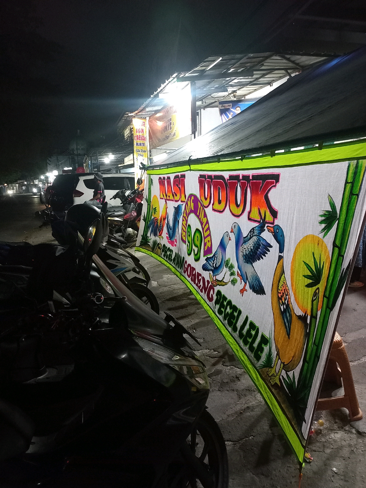 At night, a street-side food stall in Depok, Indonesia, is depicted. The foreground is dominated by several parked motorcycles. A large banner, advertising "NASI UDUK" (a type of rice dish) and "PECEL LELE" (fried catfish with chili sauce), is prominently displayed on the right side of the image. The banner features colorful illustrations of birds, a duck, and bamboo. Behind the motorcycles, a food stall structure with a corrugated metal roof is visible, illuminated by artificial lights. A white car and other motorcycles are parked further down the street, and the road appears to be paved. The overall atmosphere is that of a typical roadside eatery at night.
