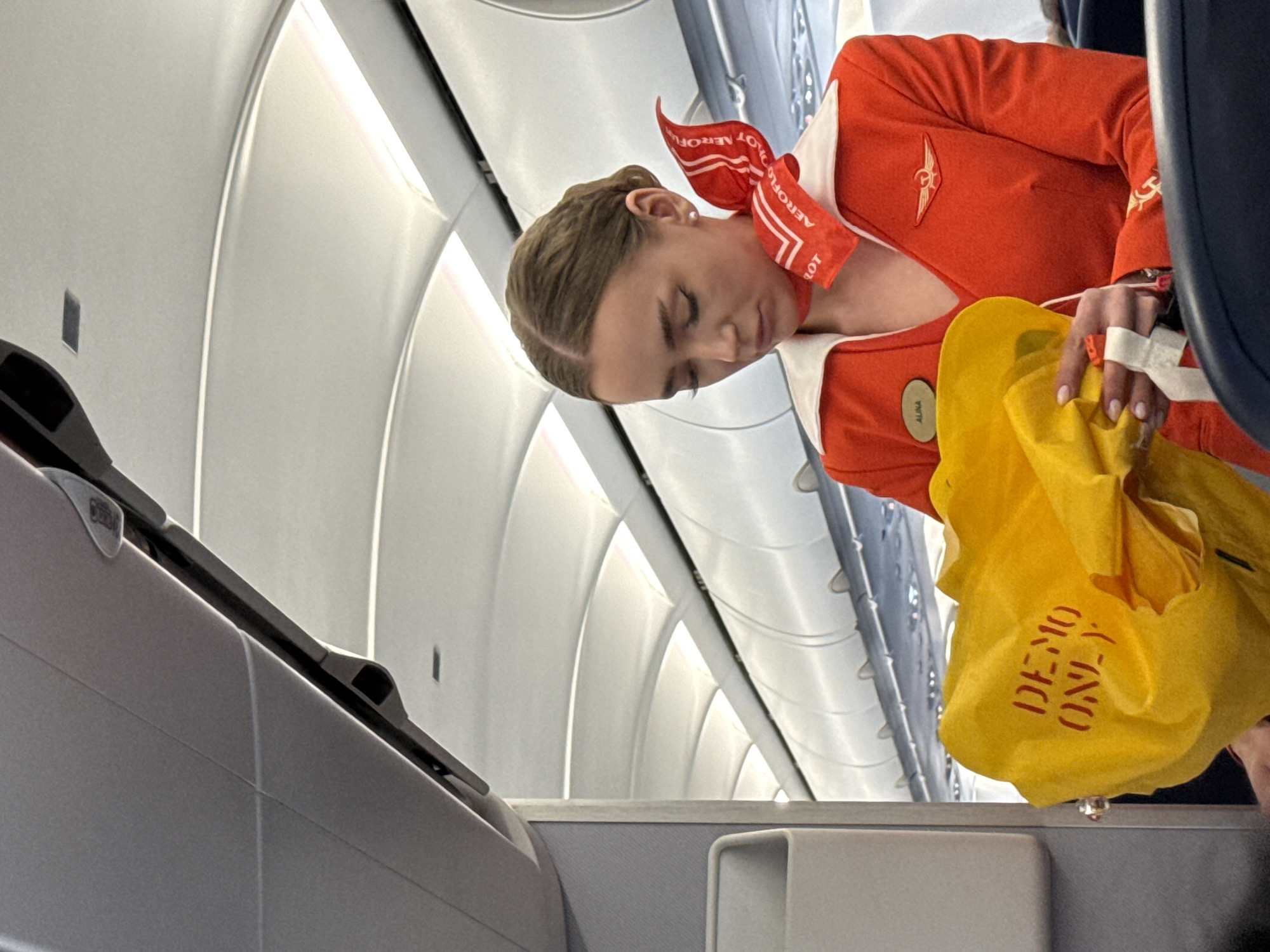 A female flight attendant, identified by the name tag "ALINA" on her orange Aeroflot uniform, is holding a bright yellow life vest in her hands inside the cabin of an airplane. The life vest has red text that reads "DEMO ONLY". She is wearing a red scarf with the Aeroflot logo. The background shows the interior of an aircraft, with curved overhead compartments and lighting. The image appears to have been taken in flight, suggesting the location is an airplane at cruising altitude, potentially over or near Khimki, Russia, as Aeroflot is the flag carrier of Russia.