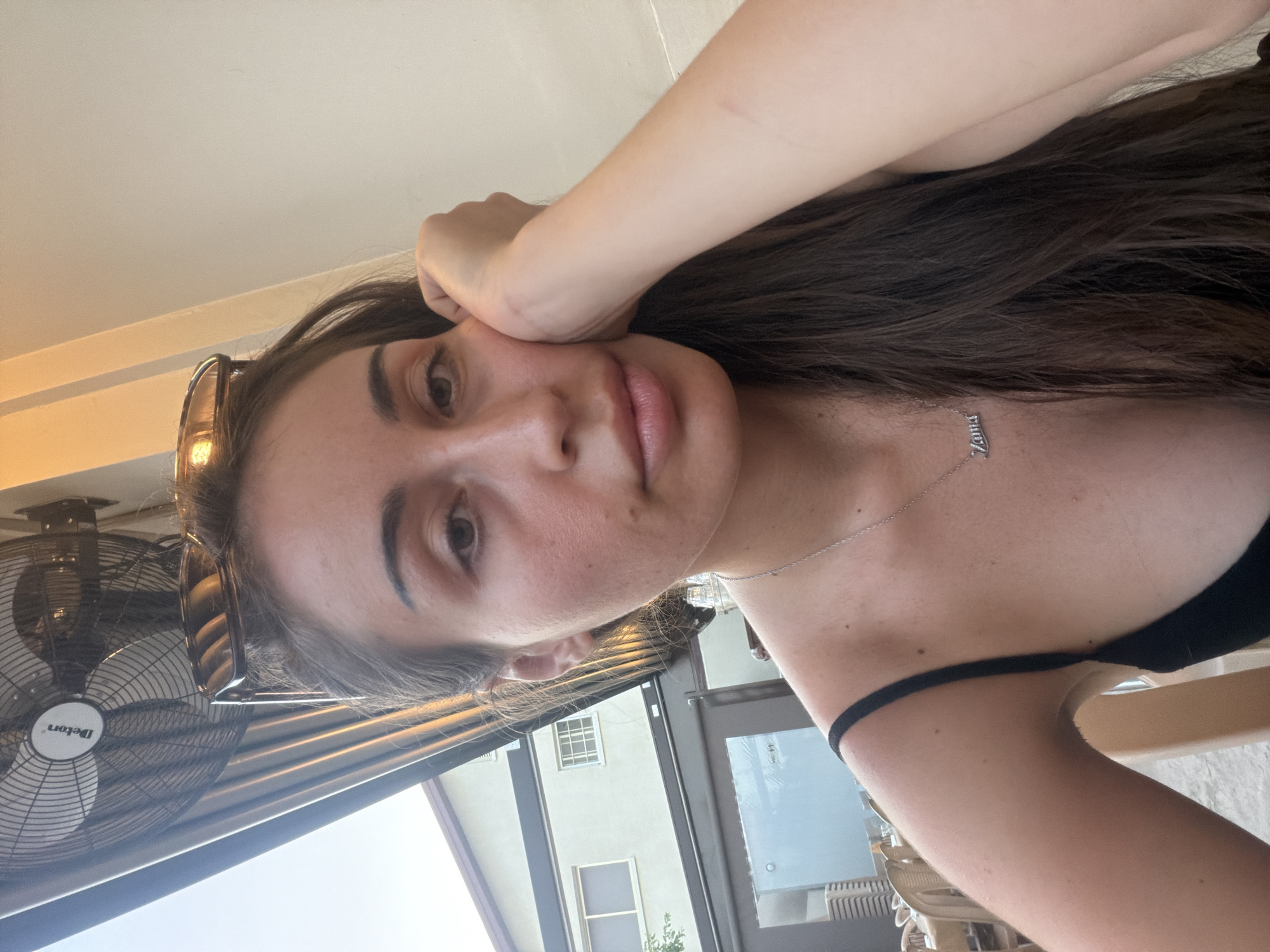 A woman is pictured in a close-up shot, resting her chin on her hand. She has long, dark brown hair and is wearing a black strap top and a silver necklace with a pendant that reads "Zama." She is outdoors, with a building and what appears to be an outdoor fan visible in the background, suggesting a cafe or restaurant setting. The lighting indicates it is daytime.