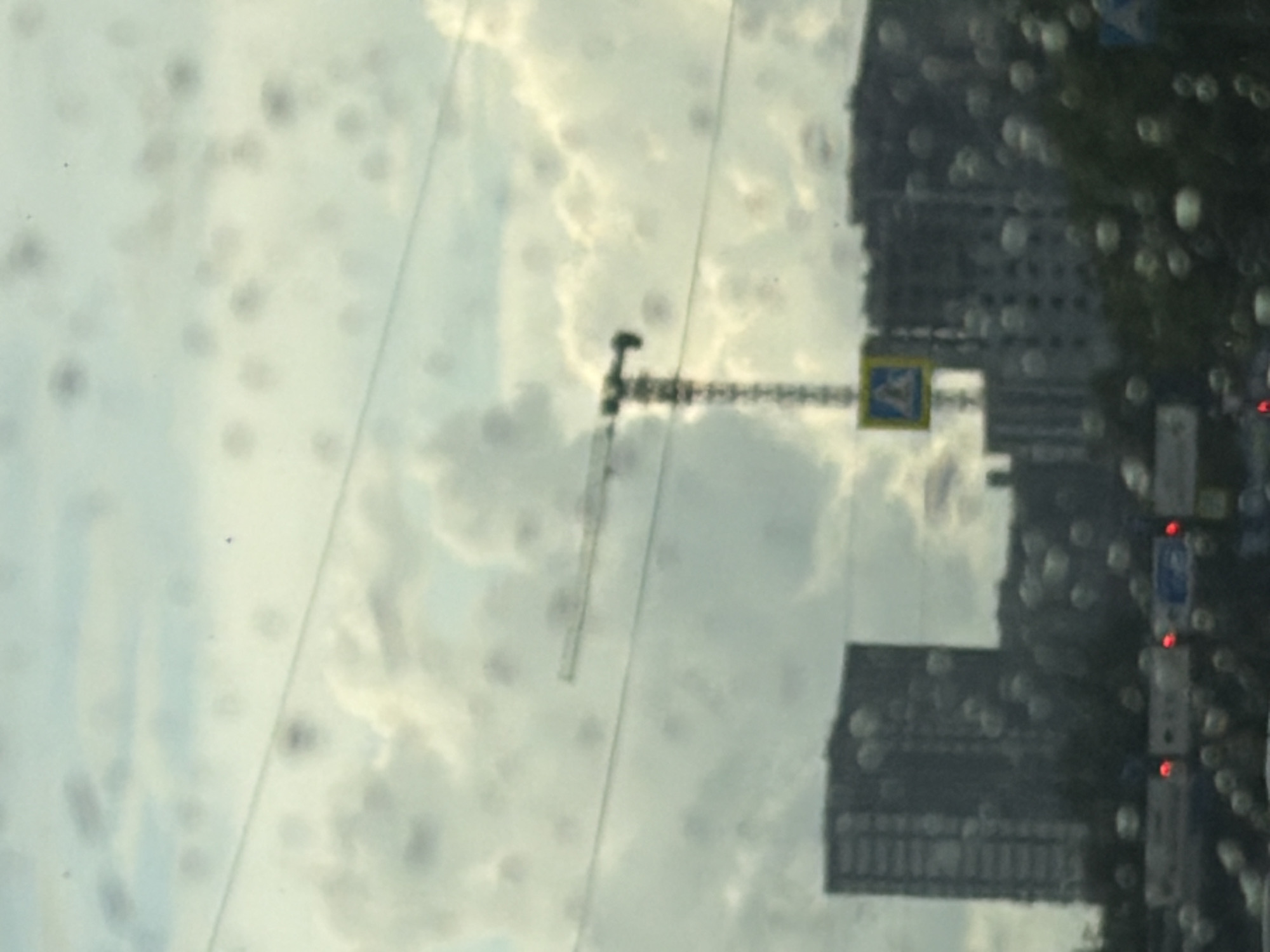The image shows a cloudy sky with a construction crane and tall buildings in the background. A yellow pedestrian crossing sign is visible on the left side of the image, below the crane. The foreground is blurred by water droplets on a surface, likely a window. The scene appears to be a city during a cloudy day.