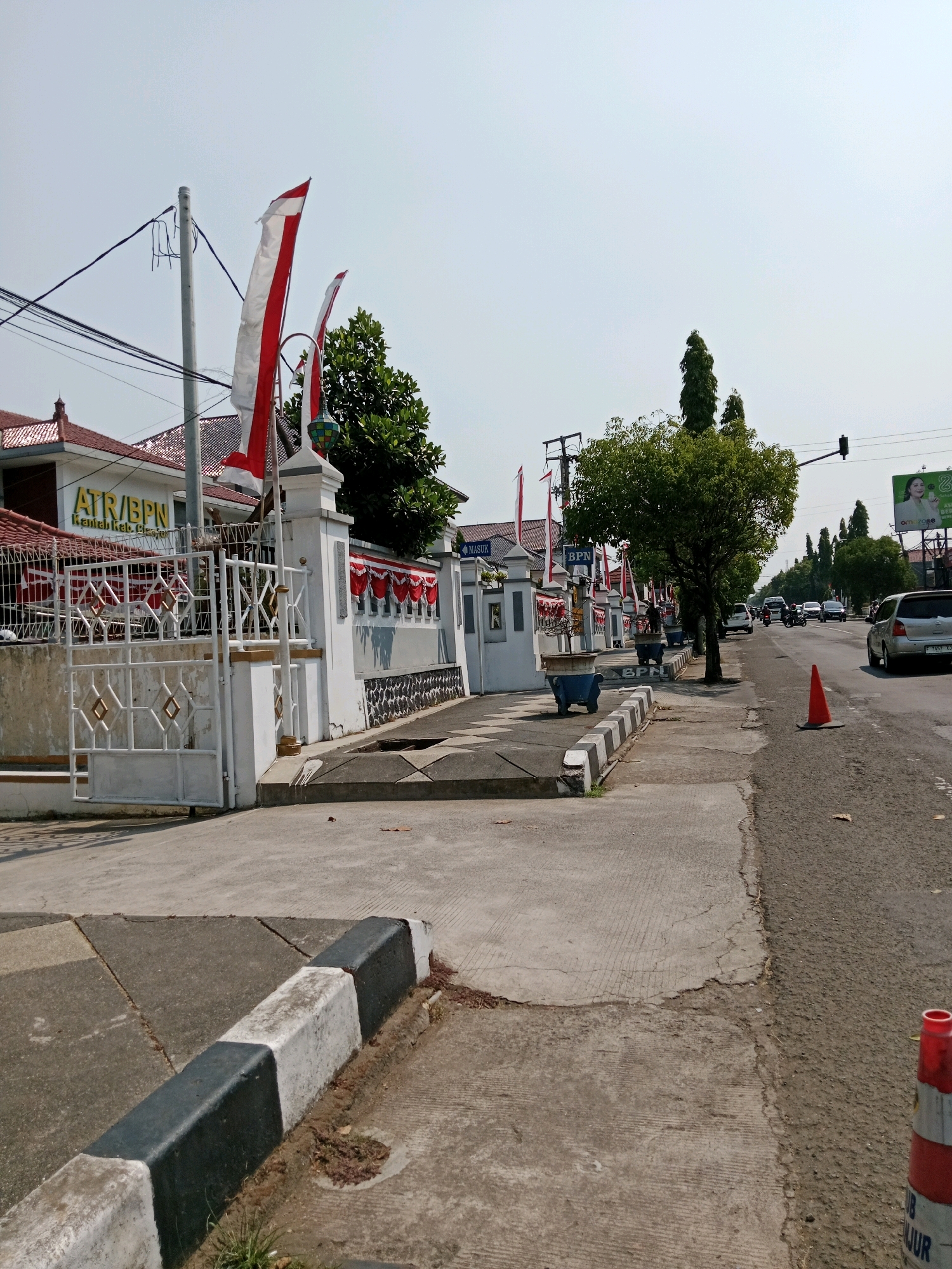 The image depicts a street scene in the city of Muka, Indonesia, likely near an official government building given the "ATR/BPN Kantor Kab. Cirebon" signage visible on the white building on the left. The building is fronted by a white metal gate and fence, adorned with red and white pennant banners, suggesting a national holiday or celebration. Several Indonesian flags, red and white, are also planted and fluttering.
The street is paved, with a sidewalk running alongside it. The sidewalk is separated from the road by a concrete curb painted with alternating black and white sections. Trees line the sidewalk, providing shade. Several cars and a motorcycle are visible on the road, suggesting moderate traffic. An orange traffic cone is placed on the right side of the road, near the curb. A large billboard is visible in the distance on the right side of the road, with a picture of a woman on it. The sky is clear and bright, indicating a daytime scene. The overall atmosphere is calm and orderly, with a touch of national pride due to the decorations.