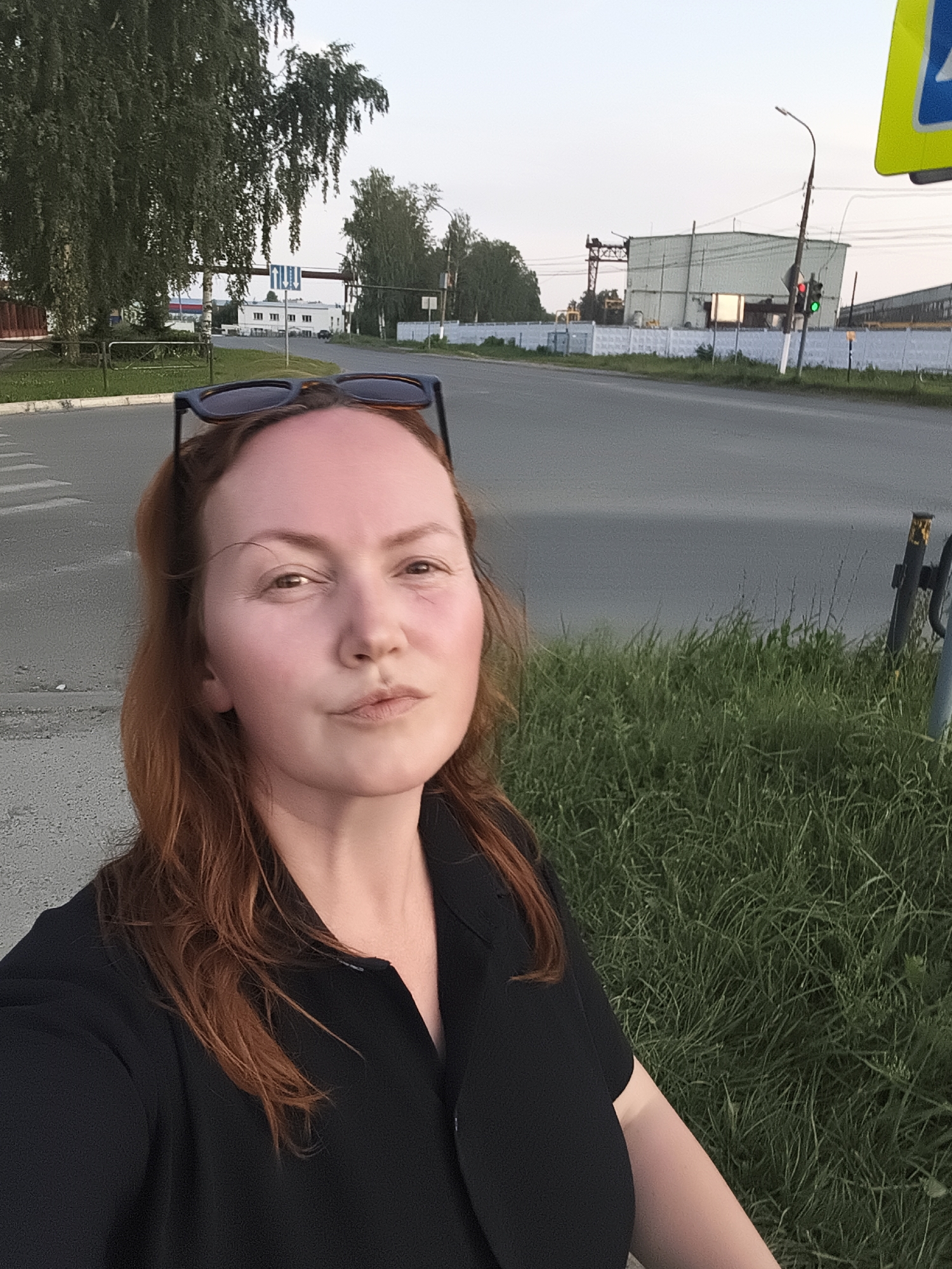 A selfie is taken by a woman with reddish-brown hair and sunglasses perched on her head. She is wearing a black collared shirt. She is standing on the side of a paved road, with a grassy area to her right. In the background, there is a traffic light, a street lamp, and industrial buildings. The scene appears to be in Chaikovsky, Russia, as indicated by the presence of a pedestrian crossing sign with Cyrillic lettering. The sky is overcast.
