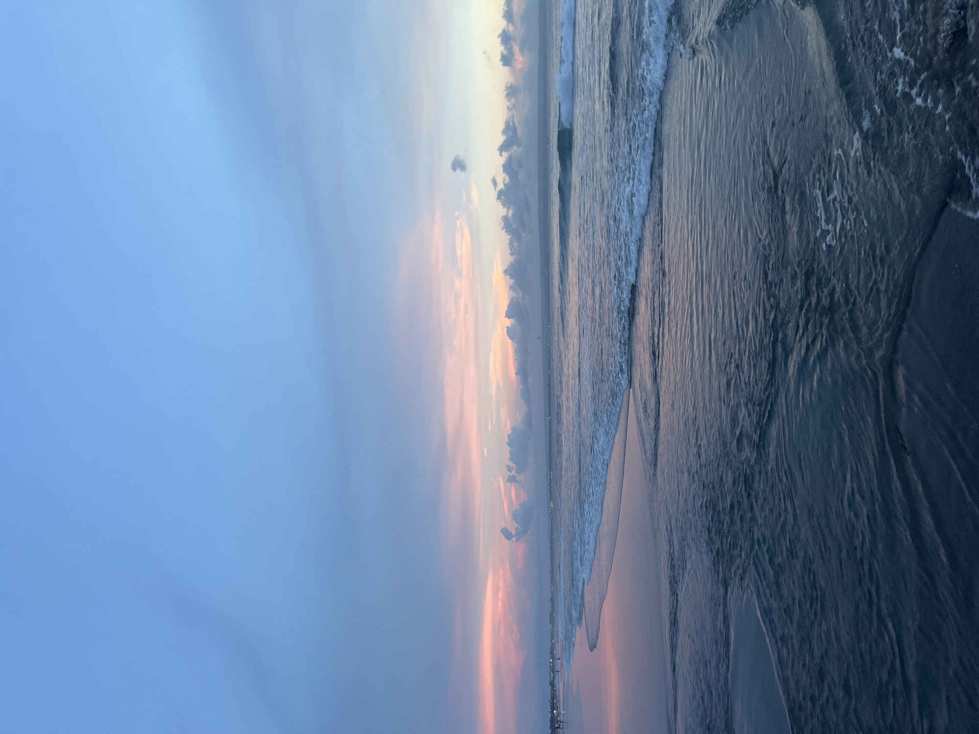 The image shows a beach scene at Kuta, Indonesia during sunset. The sky exhibits a gradient from blue to pink and orange hues, with some scattered clouds. The sea is a darker shade, with waves gently lapping against the shoreline. The sand appears dark and wet, reflecting the colors of the sky. There are people on the beach in the distance.