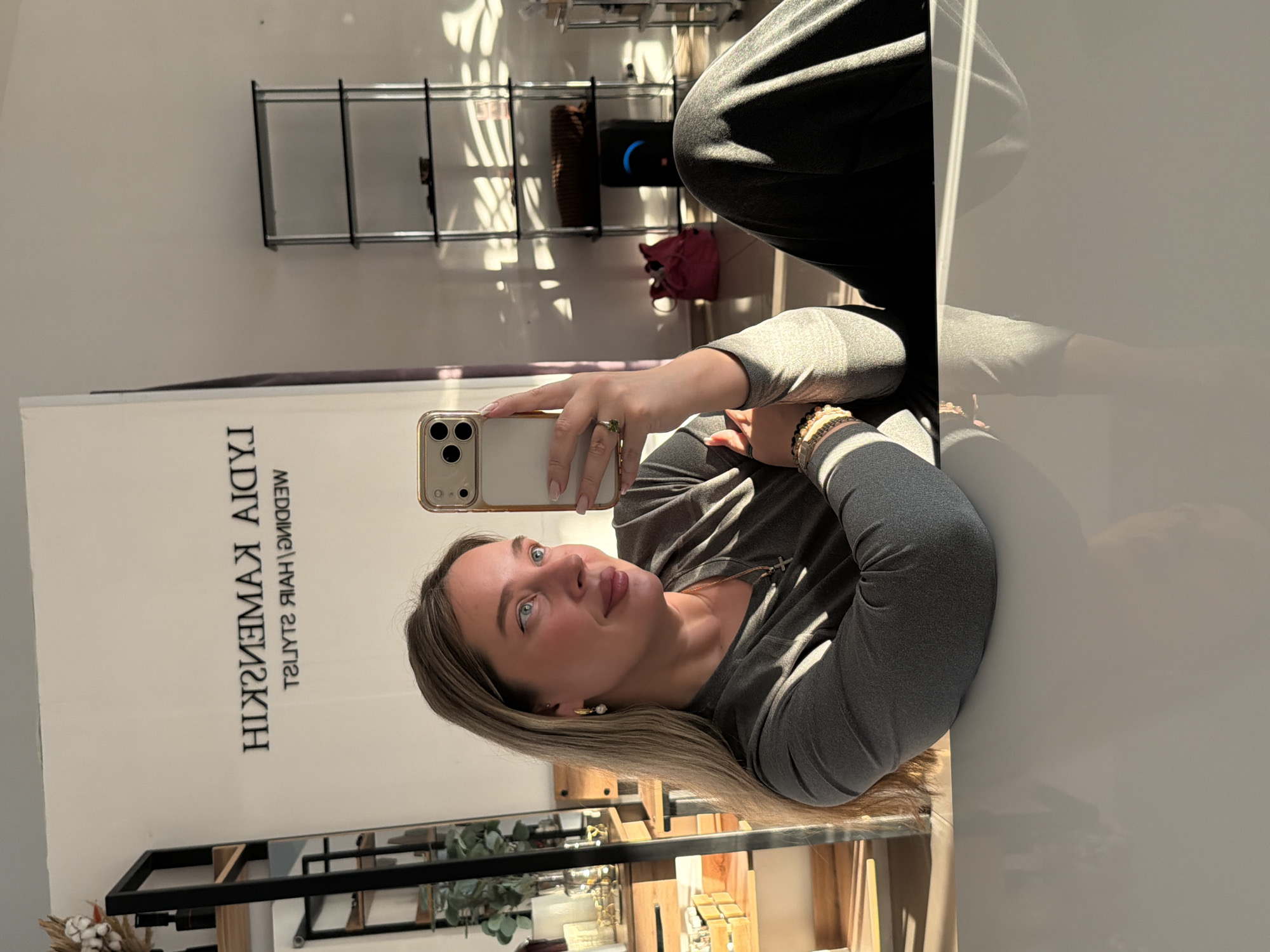 The image shows a woman taking a selfie inside what appears to be a hair salon or beauty studio. The background features a sign that reads "HIKMATSHINA TADIYA, WEDDING HAIR STYLIST". The woman is wearing a grey long-sleeved shirt and black pants. There are shelves in the background displaying various items. A red bag is visible on one of the shelves. Based on the sign, it is highly likely that the location is in Chaikovsky, Russia.
