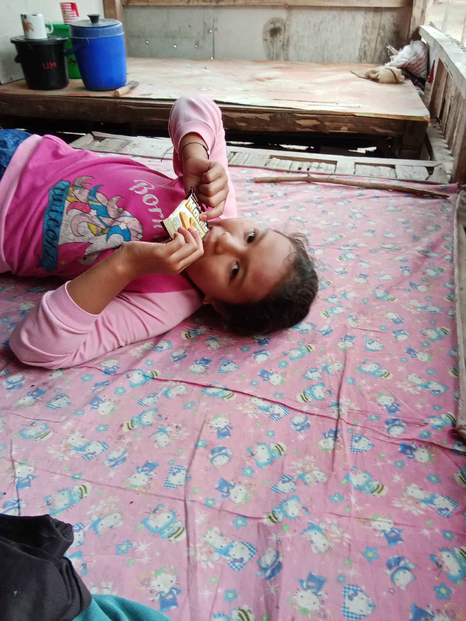 In Selowogo, Indonesia, a young girl lies on a pink bedsheet adorned with cartoon characters. She is wearing a pink shirt with a graphic print. Her right hand is holding a small, rectangular snack packet, which she is bringing towards her mouth. The background shows a rustic wooden structure with a wooden platform that appears to be a bed, and several containers, including a blue water cooler. The setting suggests a simple, possibly rural, domestic environment.