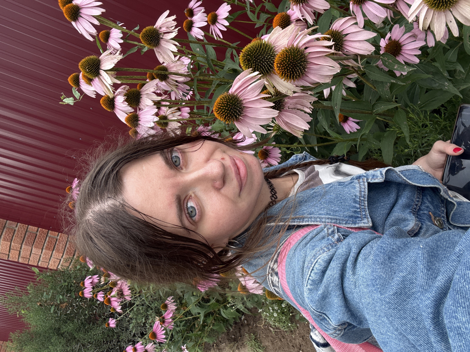A woman with blue eyes, brown hair in pigtails, and a nose ring is taking a selfie outdoors. She is wearing a denim jacket and a pink and white backpack. She is standing in front of a large patch of pink coneflower flowers, with a burgundy fence and brick wall in the background. A small green insect is visible on one of the flowers.