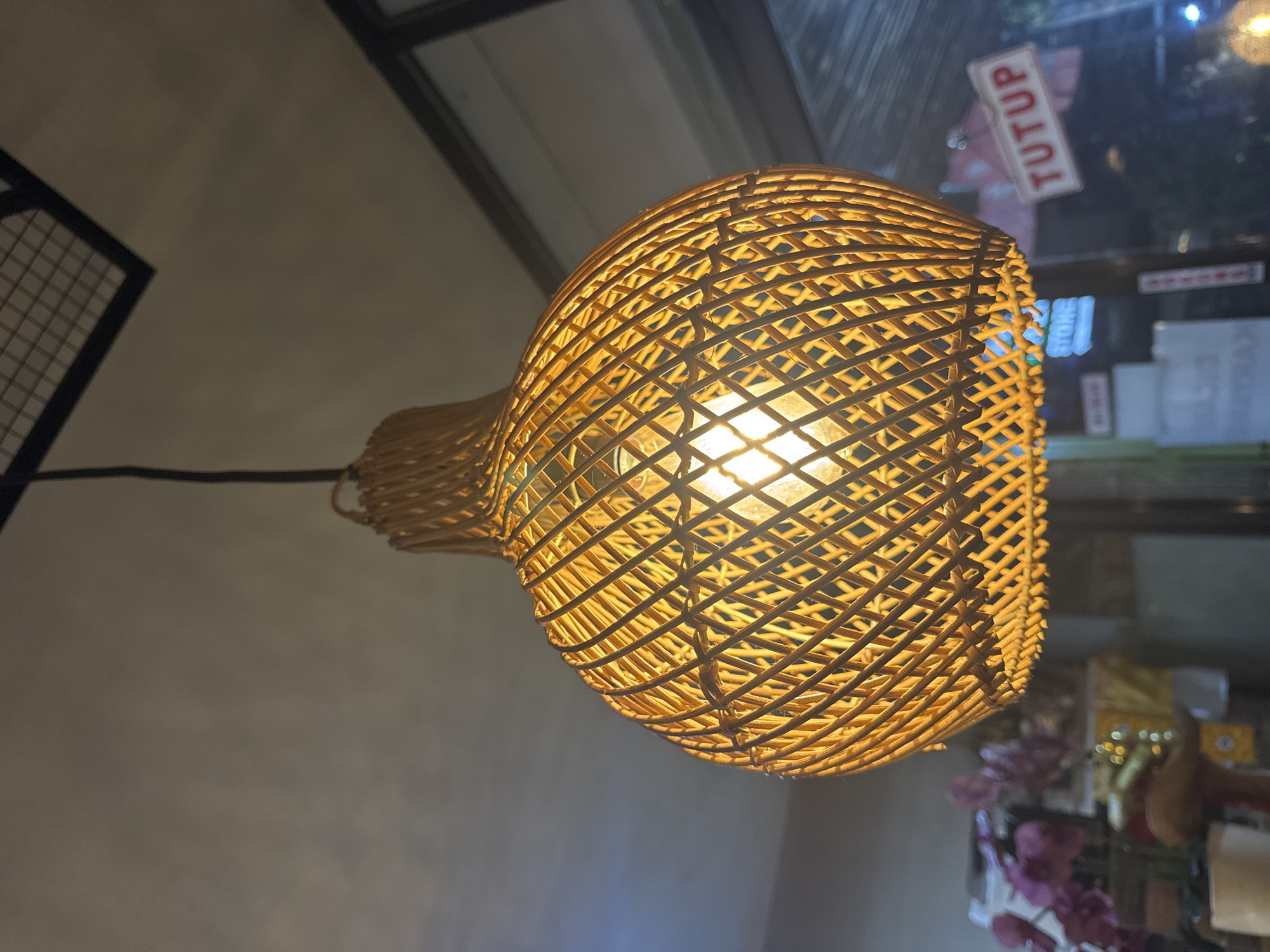 The image, appearing to be rotated 90 degrees clockwise, presents an indoor scene with a prominent, brightly lit lamp as its central subject. The lamp features a roundish shade made of light-colored woven rattan or wicker, through which the glowing light bulb is clearly visible, casting a warm, yellowish light. It hangs from the ceiling, suggesting a commercial interior space.
Through a window or glass door in the background, a white rectangular sign with the word "TUTUP" written in red letters is visible. "TUTUP" is Indonesian for "CLOSED," providing a strong location cue consistent with Kuta Selatan, Indonesia. The exterior beyond the window appears dark, illuminated by artificial lights, indicating the photograph was likely taken at night or dusk. Blurred reflections of other outdoor lights and structures are also present.
In the upper left portion of the image, a dark, grid-like structure is partially visible, possibly part of a ceiling or architectural detail. Below the lamp on the right, some out-of-focus elements, including what looks like purple or pink decorative flowers, contribute to the interior setting. The overall atmosphere conveyed by the warm lighting is cozy and inviting. There are no people visible, and no specific activity is taking place.