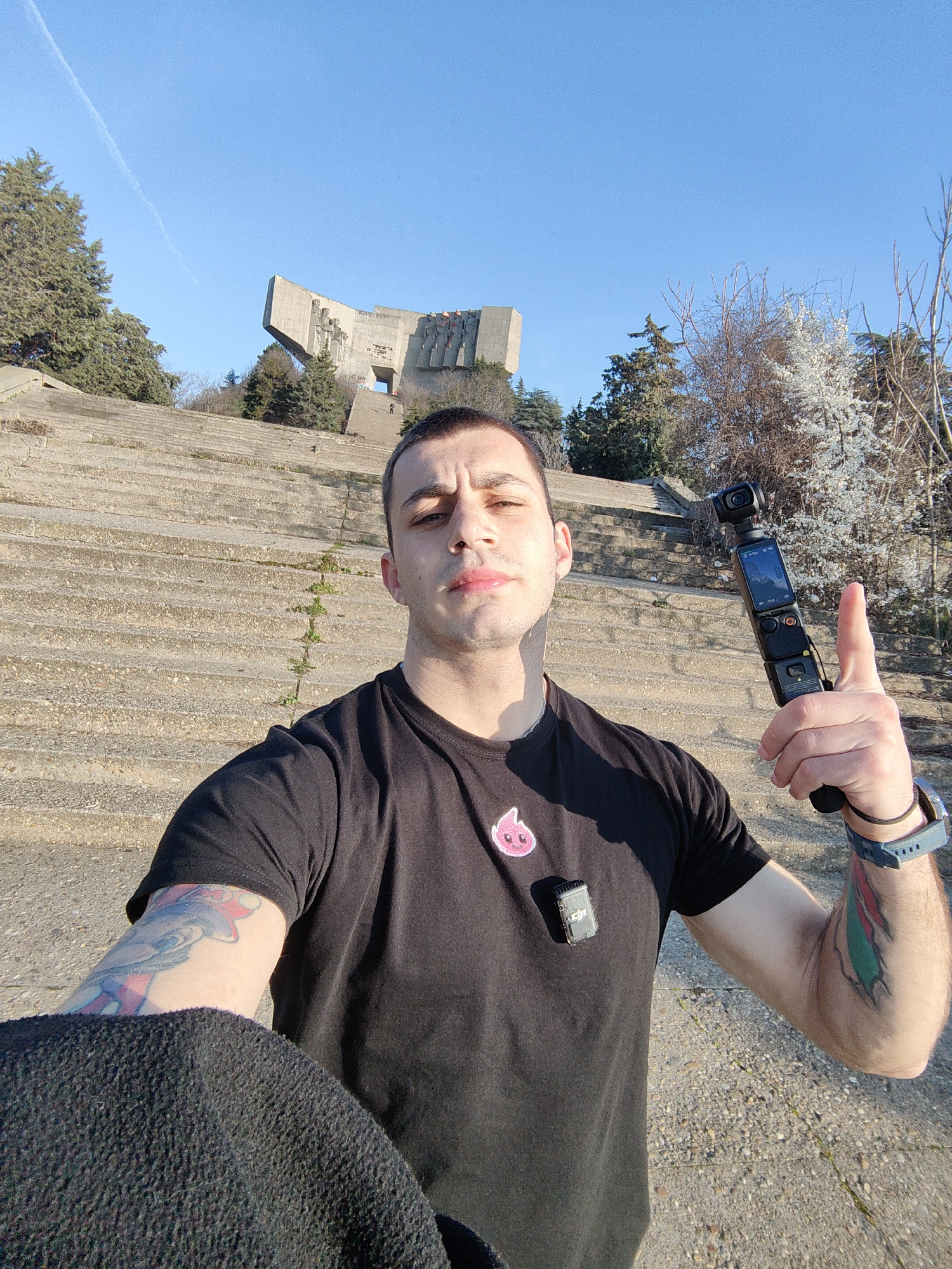 A male individual is depicted in a selfie photograph taken outdoors at the Monument of Bulgarian-Soviet Friendship in Varna, Bulgaria. The subject, a man in his 20s or 30s with short dark hair, wears a black short-sleeved t-shirt featuring a pink, cartoonish, drop-shaped emblem on the left chest. A grey DJI Mic wireless microphone transmitter is clipped to his shirt. He exhibits visible tattoos: a colorful character on his left forearm and a multi-colored abstract design on his right forearm. A grey-blue digital watch is worn on his left wrist. He holds a black DJI Osmo Pocket 3 camera in his right hand, extended forward towards the viewer, with his thumb raised in a 'thumbs up' gesture.
The background features the Monument of Bulgarian-Soviet Friendship, a large, brutalist-style concrete structure, situated at the apex of a broad, multi-tiered concrete staircase. The steps show wear and minor vegetation growth in cracks. Evergreen trees and deciduous trees, some with early white blossoms, flank the monument and stairs. A faint human figure is visible on the upper section of the monument's steps. The sky is clear and bright blue with a single, thin contrail. The scene is well-lit by direct sunlight, suggesting a clear day.