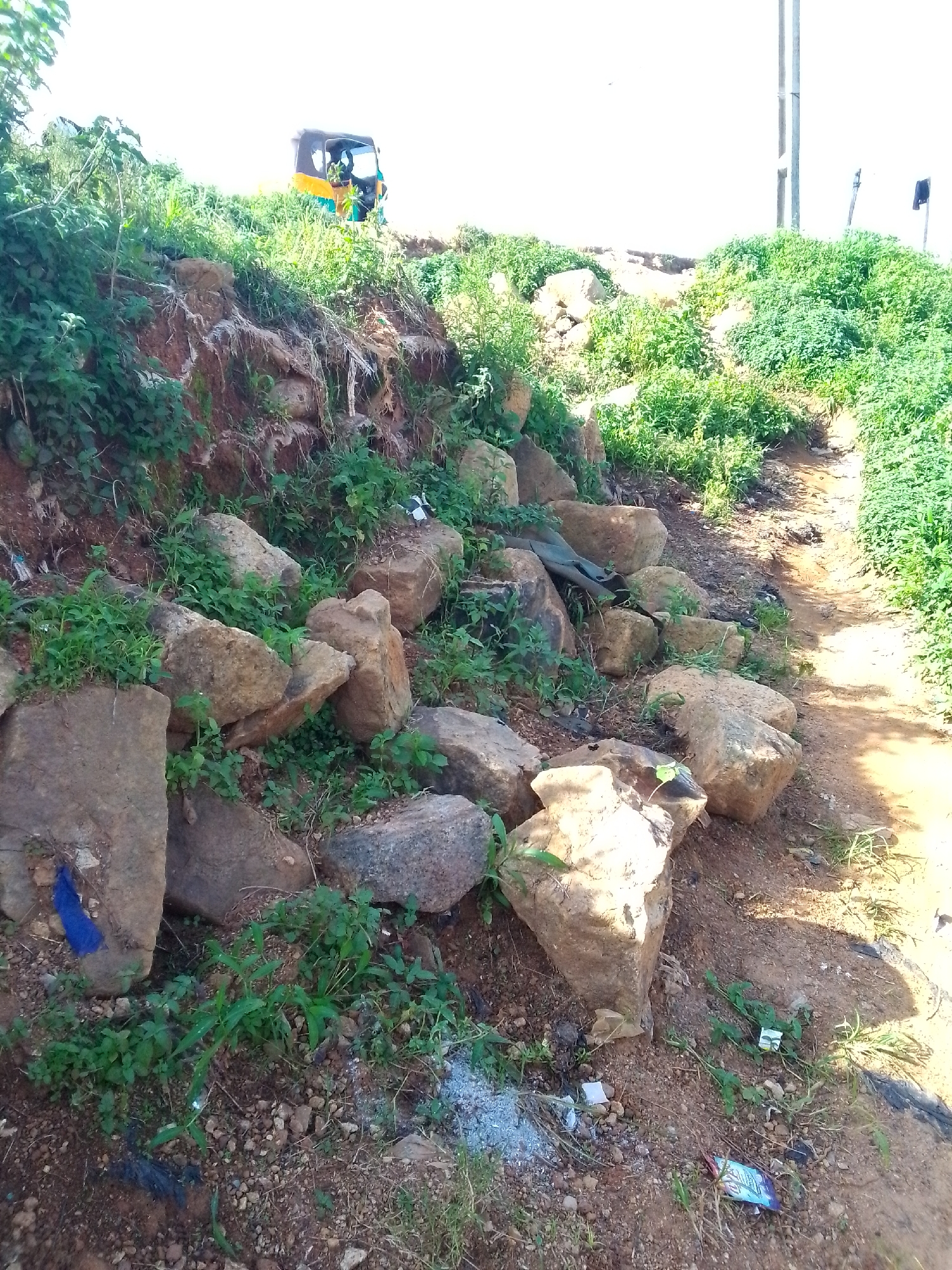 An eye-level, medium shot captures a roadside embankment with scattered rocks and vegetation in Jos, Nigeria. A yellow and blue auto-rickshaw, likely a "Keke Napep," is visible on the top of the embankment, partially obscured by greenery. The embankment is characterized by exposed reddish-brown soil and a variety of rocks, ranging from small pebbles to larger boulders. Lush green grass and other plants grow around and between the rocks, indicating a potentially untended or natural area. A dirt path runs alongside the embankment on the right side of the frame. In the background, a utility pole with wires and a small dark sign are present. The ground is littered with small pieces of debris, including what appears to be plastic wrappers and small pieces of white material.