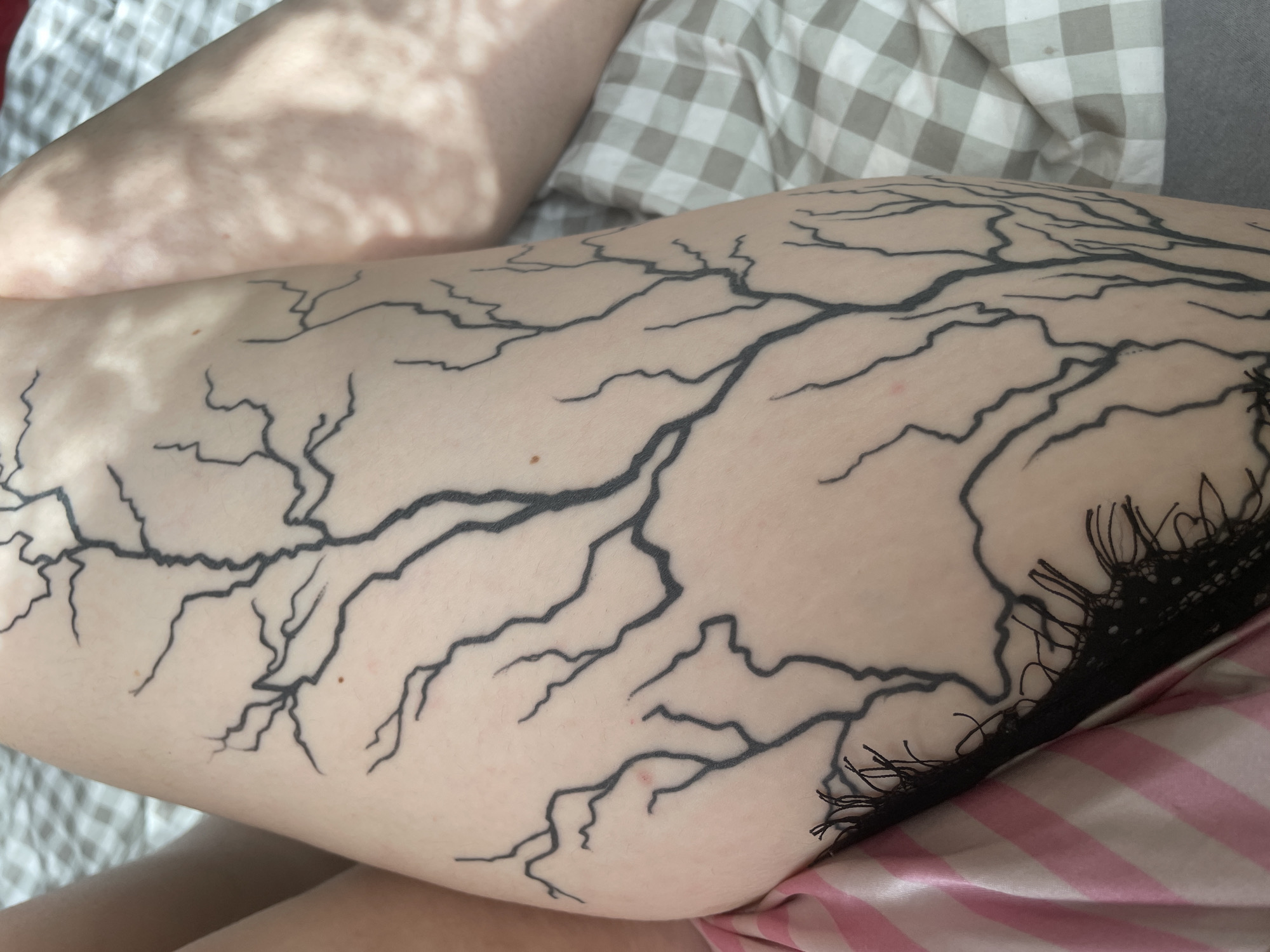 The image shows a close-up view of a tattoo on a person's thigh. The tattoo depicts a stylized tree with thin, branching lines that cover a significant portion of the thigh. The person is wearing pink and white striped satin shorts with black lace trim. The background consists of a checkered blanket. There is no visible information to indicate the location in the city of Alūksne, Latvia. The lighting suggests an indoor setting, possibly a bedroom, with natural light casting shadows on the person's leg.