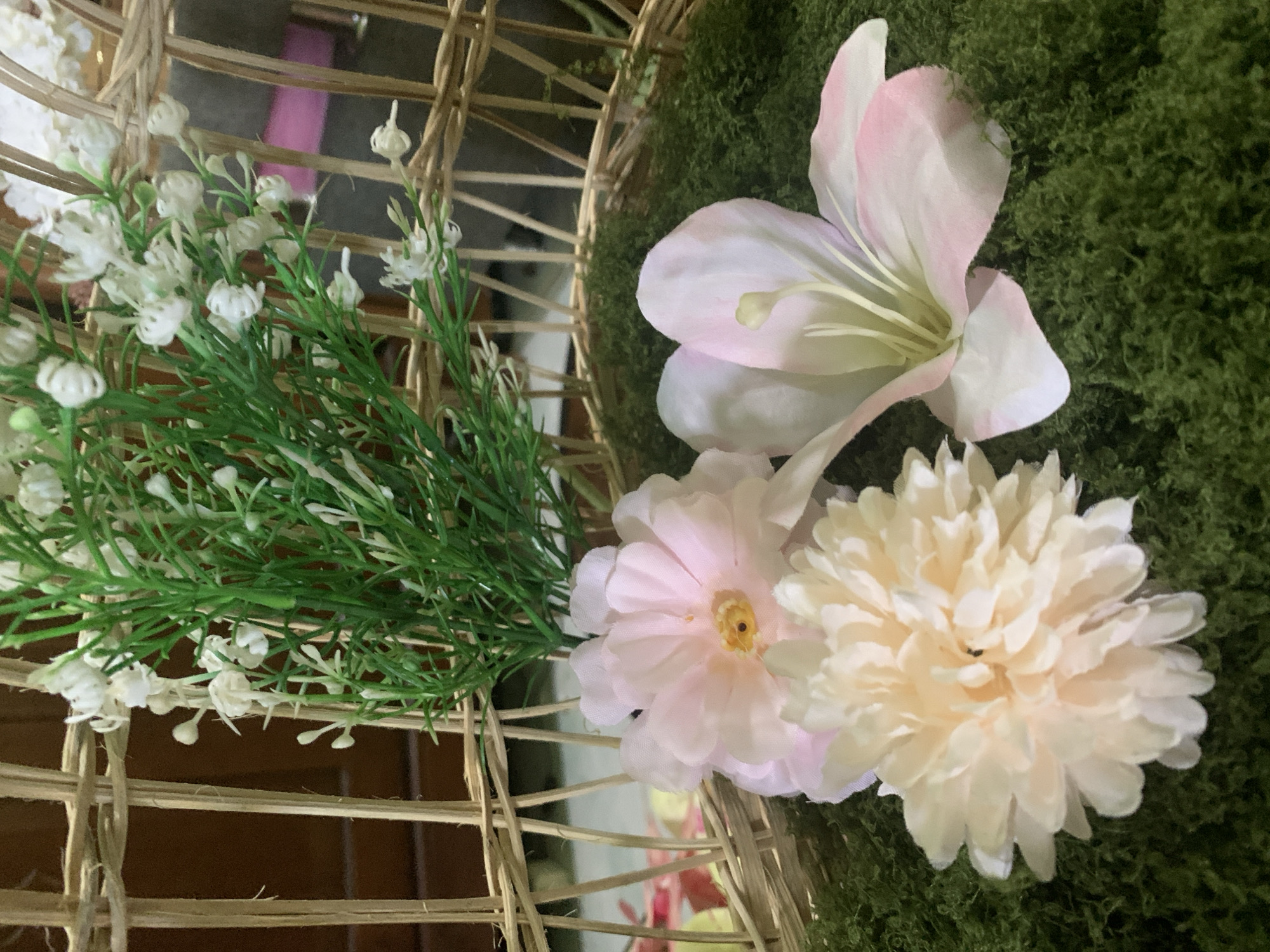 The image shows a close-up of artificial flowers arranged in a wicker basket. The basket is filled with green moss, and several pastel-colored artificial flowers are placed on top of the moss. One flower is a light pink, with a yellow center and ruffled petals. Another is a pale cream color, with numerous thin petals creating a full, round bloom. A third flower is a light pink lily, with prominent light yellow stamens. Behind the flowers, a bunch of artificial green foliage with small white flowers is visible, also contained within the basket. The wicker basket has a lattice pattern. The background is blurred but suggests an indoor setting with wooden furniture. There is no indication of the location being in Kuala Pembuang, Indonesia.