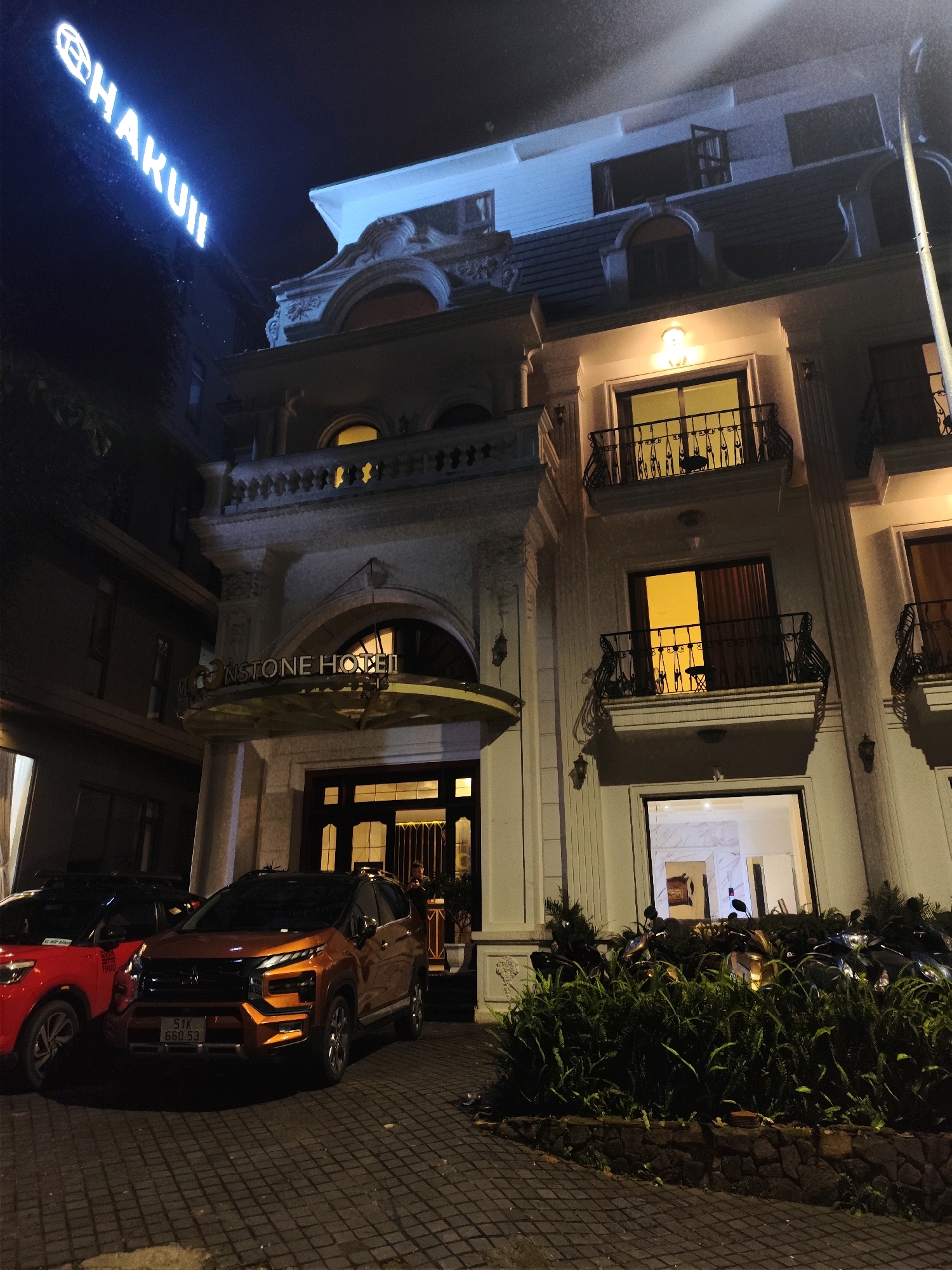 The image shows the entrance of the "STONE HOTEL" in Da Lat, Vietnam, at night. A bright blue neon sign reading "HAKUII" is visible on the left side of the building, illuminated against the dark sky. The hotel facade is made of light-colored stone with ornate architectural details, including balconies with black metal railings and arched windows. Several windows are lit from within, casting a warm yellow glow. Two cars are parked in front of the hotel entrance: a red car on the left and an orange SUV on the right. The SUV has a license plate reading "51K 660.53". The ground in front of the hotel is paved with dark interlocking bricks. A dense arrangement of green foliage and shrubs is located to the right of the entrance, with several motorcycles parked behind it, partially obscured by the plants. The overall scene suggests a well-lit, established hotel in a city environment at nighttime.