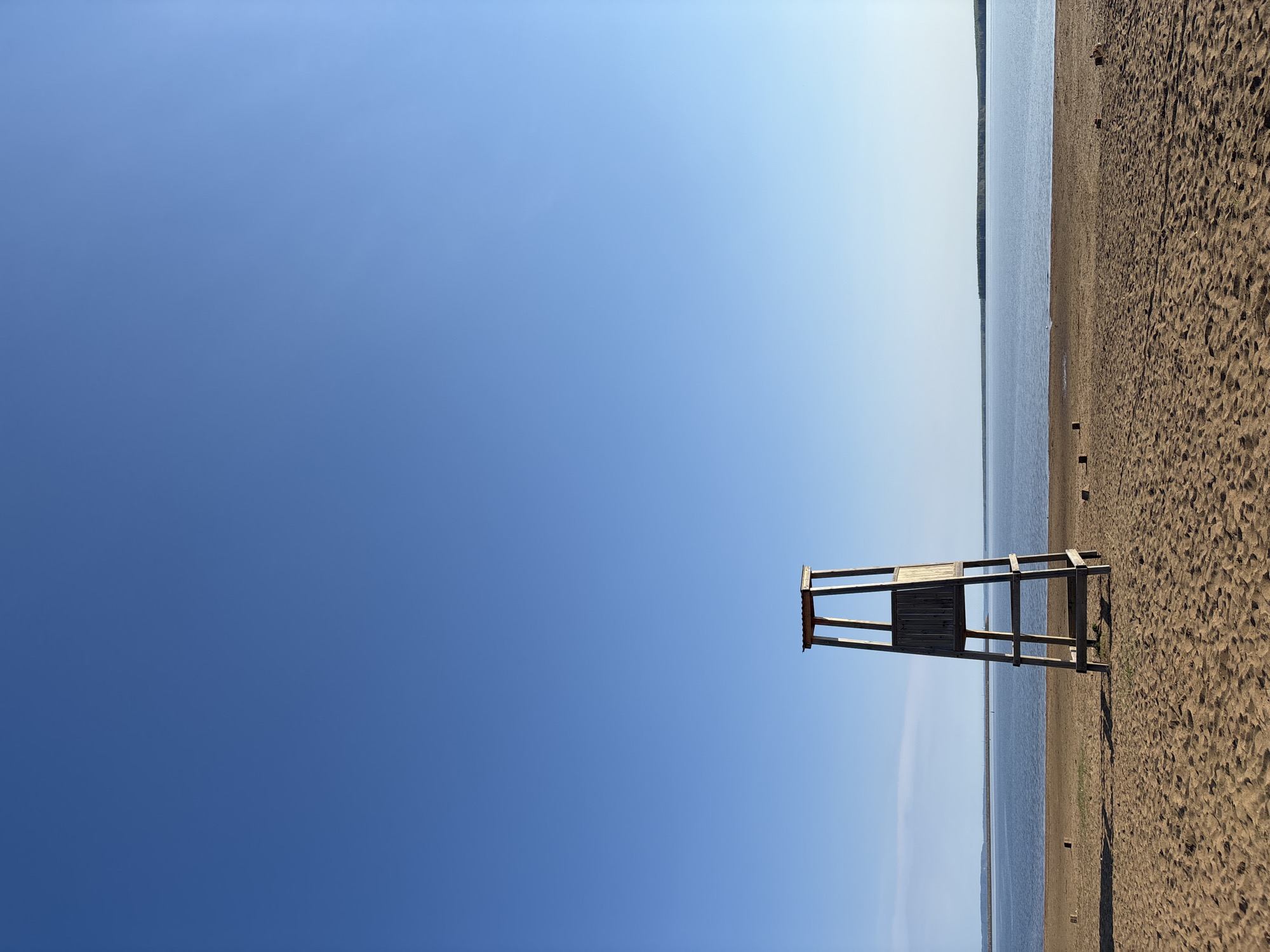 A lifeguard chair stands on a sandy beach in Chaikovsky, Russia. The sky is clear and blue. Water is visible behind the chair.