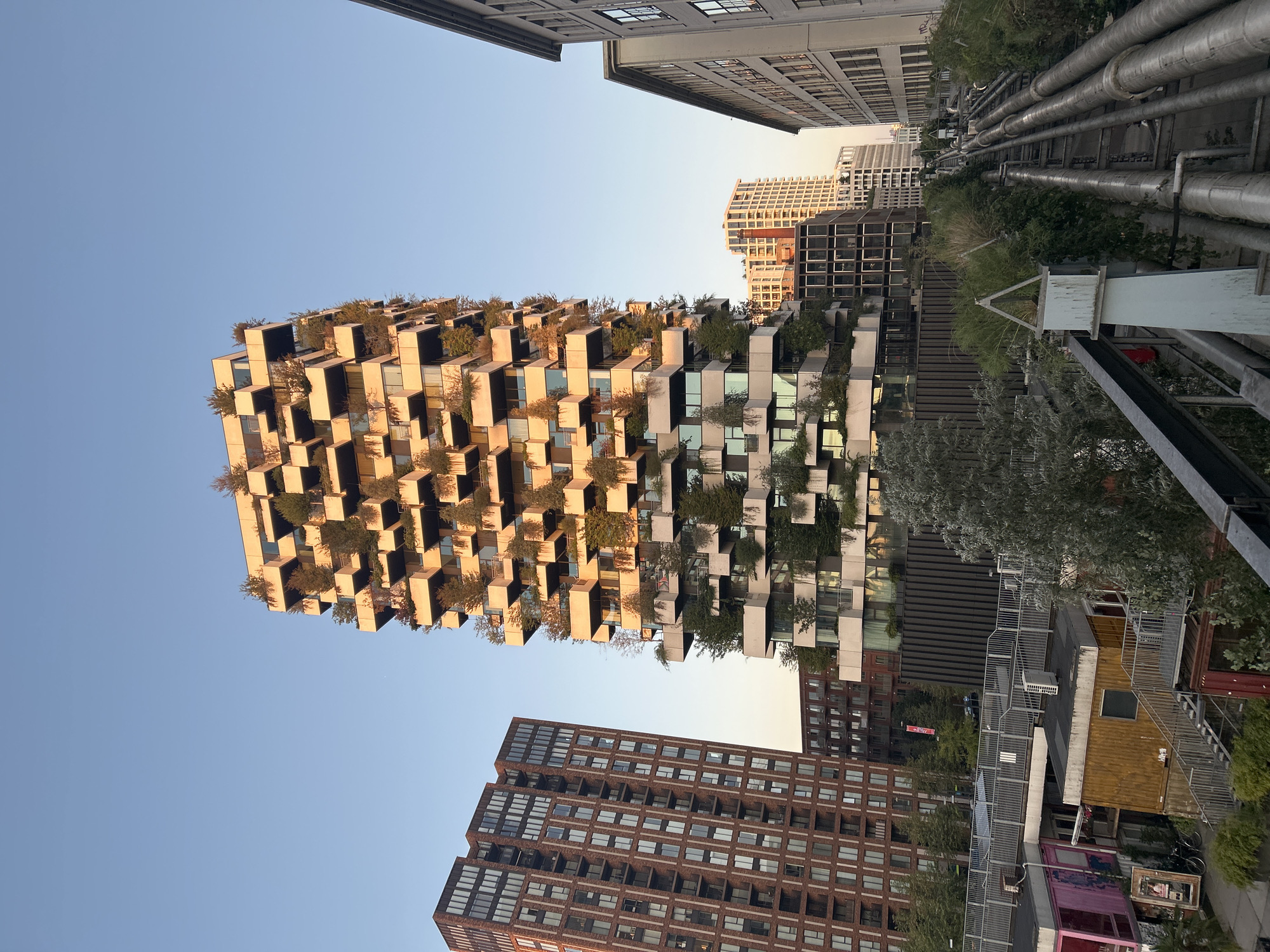 The image shows the "Wall Houses" building, designed by architect Winy Maas, located in Eindhoven, Netherlands. The building is characterized by its unique, cantilevered cube-shaped apartments, each adorned with trees and greenery. In the foreground, a raised walkway with industrial pipes running along its side suggests an urban infrastructure setting. Other buildings, including a tall red brick apartment complex and modern office buildings, are visible in the background, contributing to a dense urban landscape. The clear blue sky indicates it is daytime, possibly late afternoon given the warm lighting on the buildings. There are no people visible in the image.
