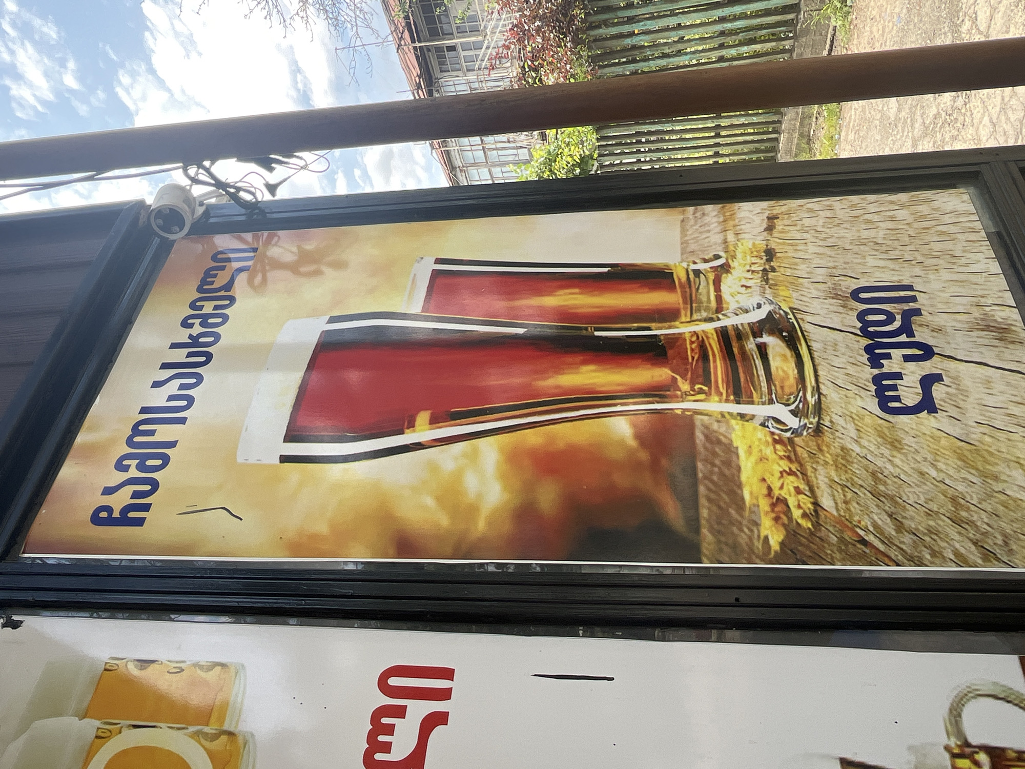The image displays a vertical poster advertising beer, written in Georgian script. The poster features two tall glasses filled with dark amber liquid, likely beer, set against a warm, glowing background. There are also stalks of wheat scattered on a wooden surface in the foreground. To the left of this poster, another poster is partially visible, showing the top of two yellow-colored beverages in clear containers. A security camera is mounted on a pole to the right of the beer poster. In the background, beyond a rustic wooden fence, a building with multiple windows and a staircase is visible, along with trees and foliage. The scene appears to be outdoors, likely on a street or sidewalk. The sign itself is framed in black. The location is identified as Surami, Georgia.