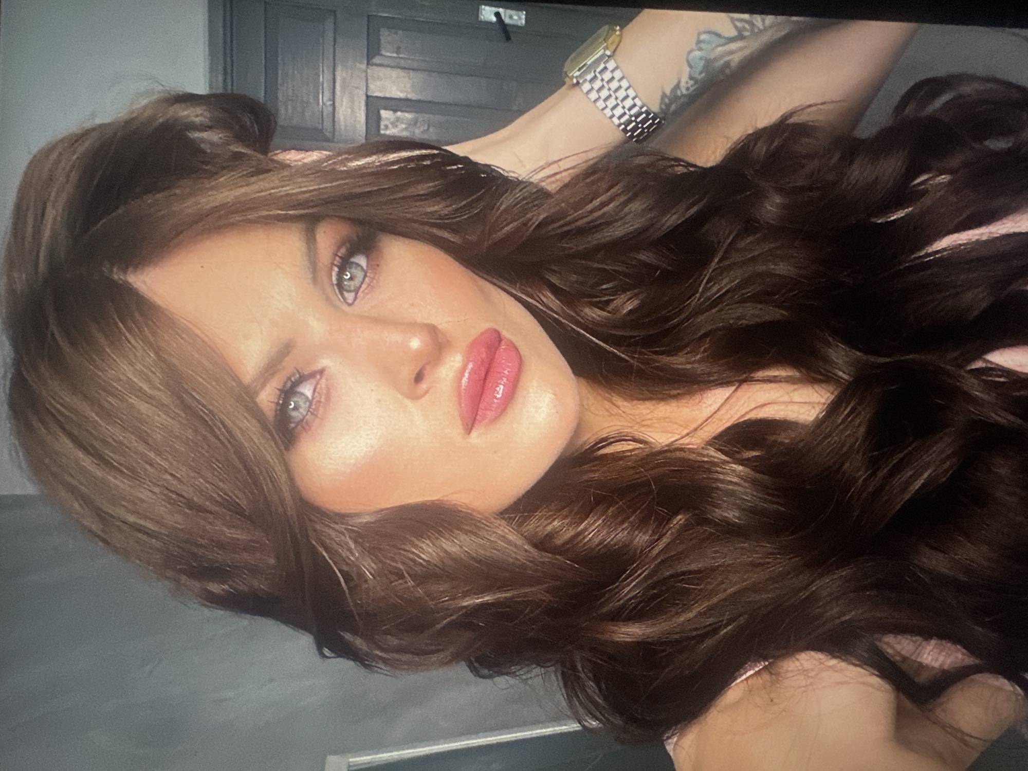 A woman with long, dark, wavy hair and blue eyes is looking directly at the camera. She has full, pink lips and is wearing makeup. She is wearing a light pink top and a silver watch on her left wrist. She has tattoos on her left arm. The background appears to be indoors, with a grey wall on the left and a dark wooden door on the right. The lighting is bright, casting highlights on her face and hair.